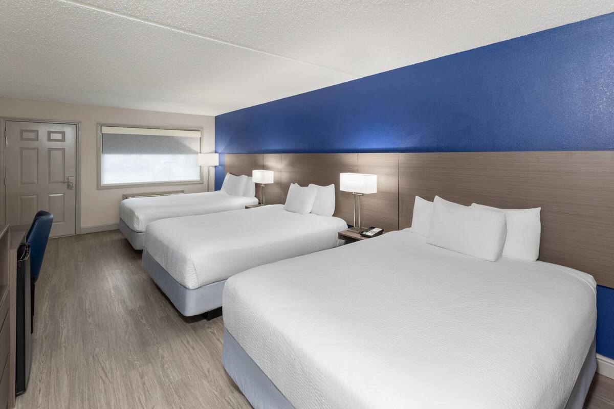 A hotel room with two beds, a desk, a chair, a window, and a blue accent wall.