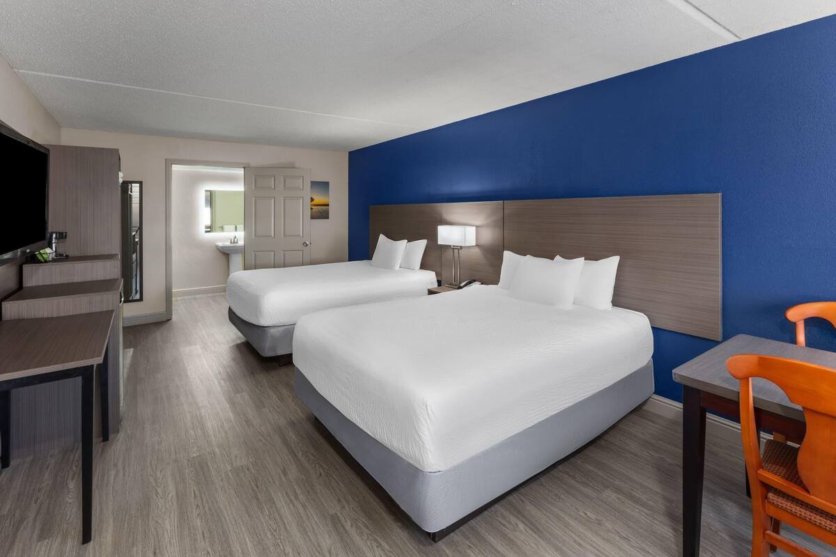 A hotel room with two beds, a desk, a chair, a TV, and a bathroom visible through an open door.