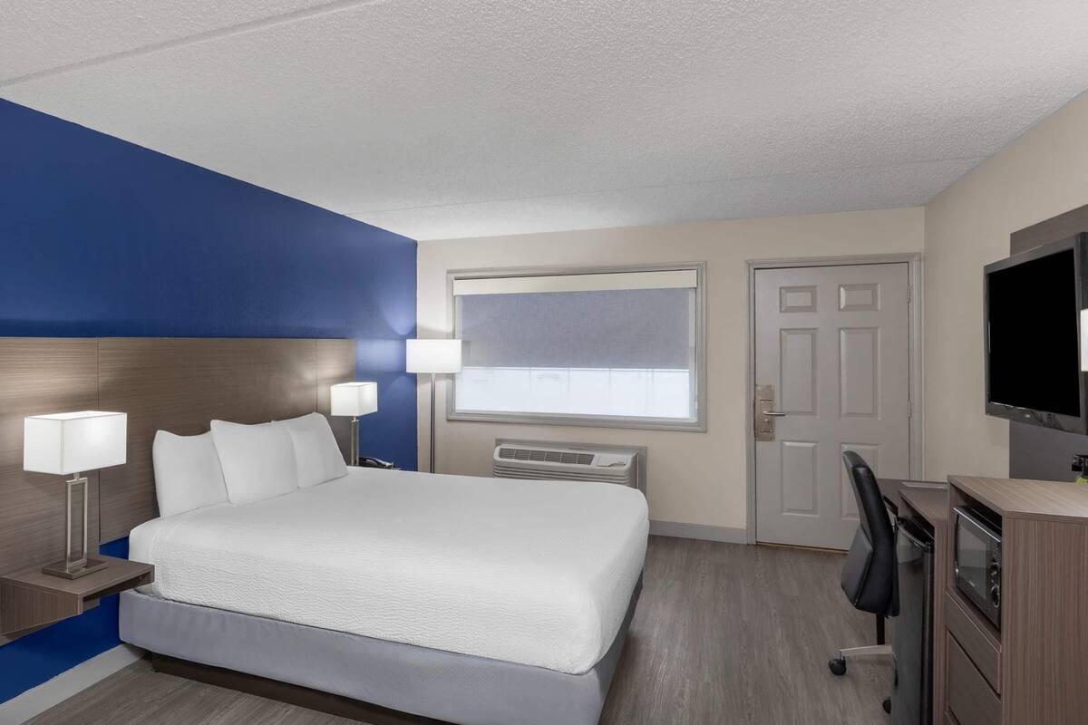 A hotel room with a bed, bedside lamps, a desk with a chair, a television, and a window with blinds.