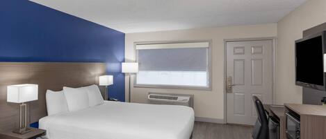 Quarto de hotel com cama, abajures, escrivaninha com cadeira, televisão e janela com persianas.