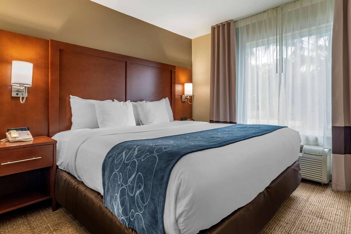 Habitación de hotel con una cama grande, cabecera de madera, lámparas de noche, un teléfono y una ventana con cortinas.