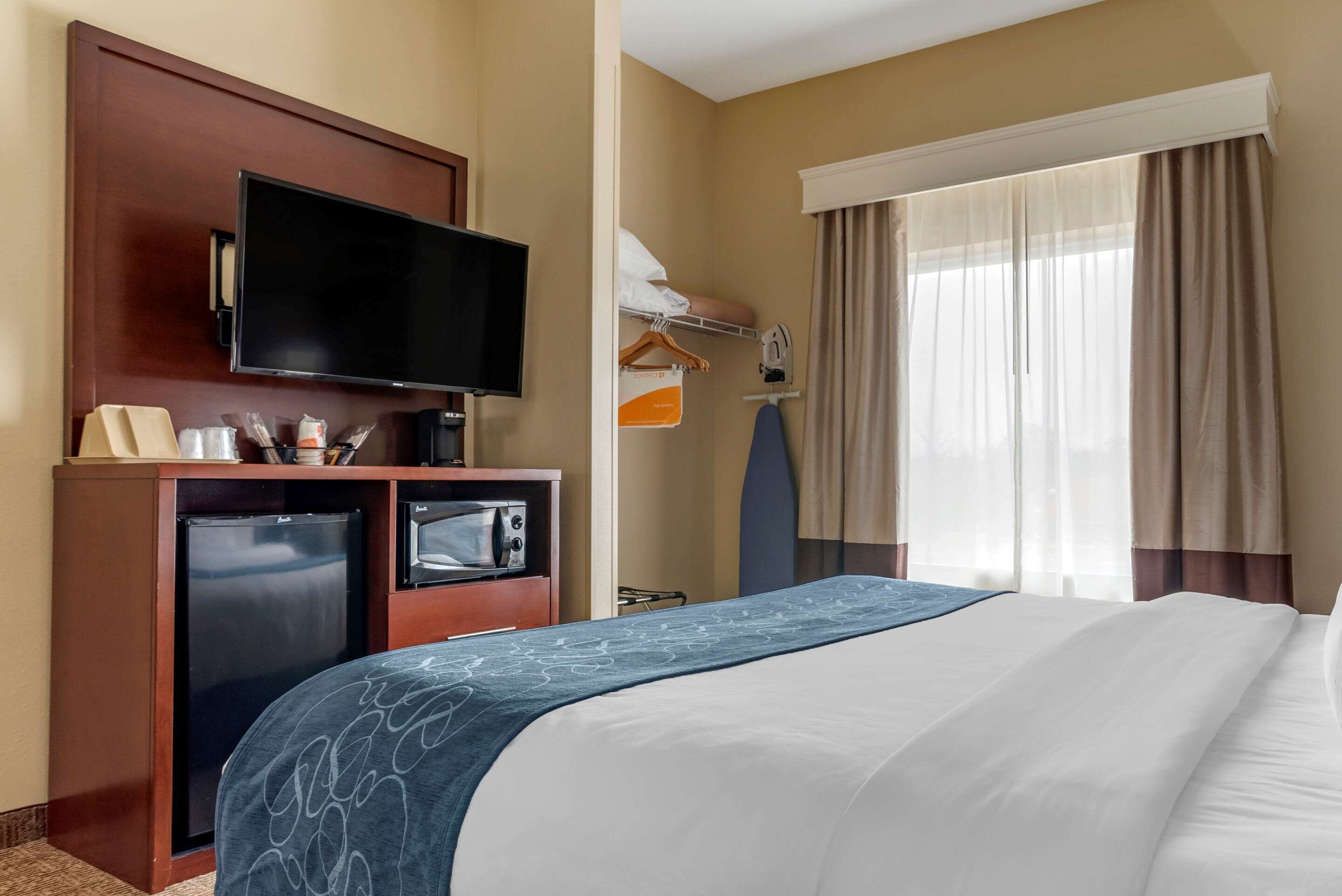 A hotel room with a bed, a television, a mini-fridge, a microwave, a desk with a computer, and a window with curtains.