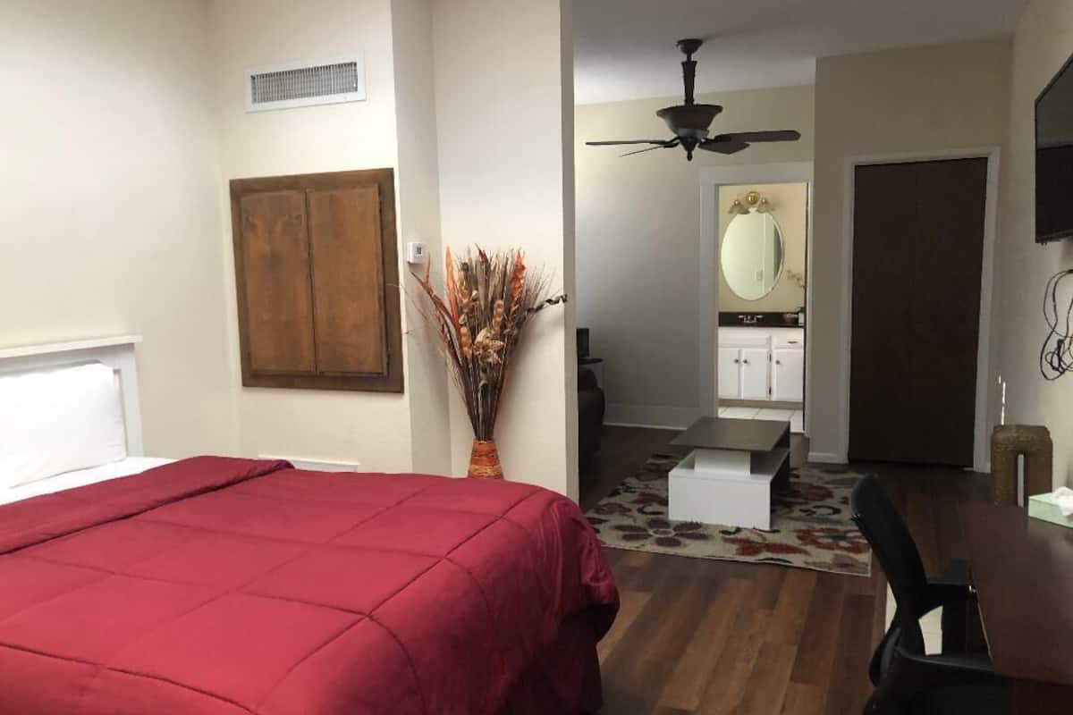 A bedroom with a bed, a wooden wardrobe, a ceiling fan, a bathroom with a mirror, and a dining area with a table and chairs.