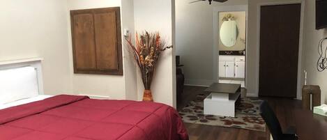Un dormitorio con cama, un armario de madera, ventilador de techo, baño con espejo y zona de comedor con mesa y sillas.