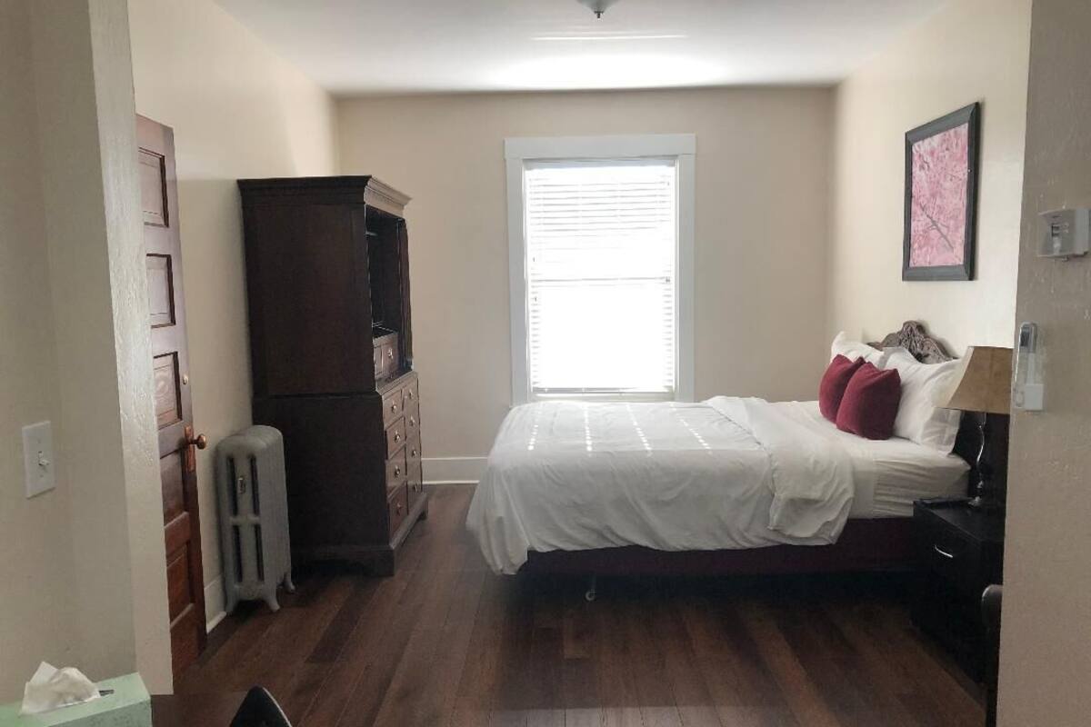 A bedroom with a bed, a dresser, a radiator, a window with blinds, and a framed picture on the wall.