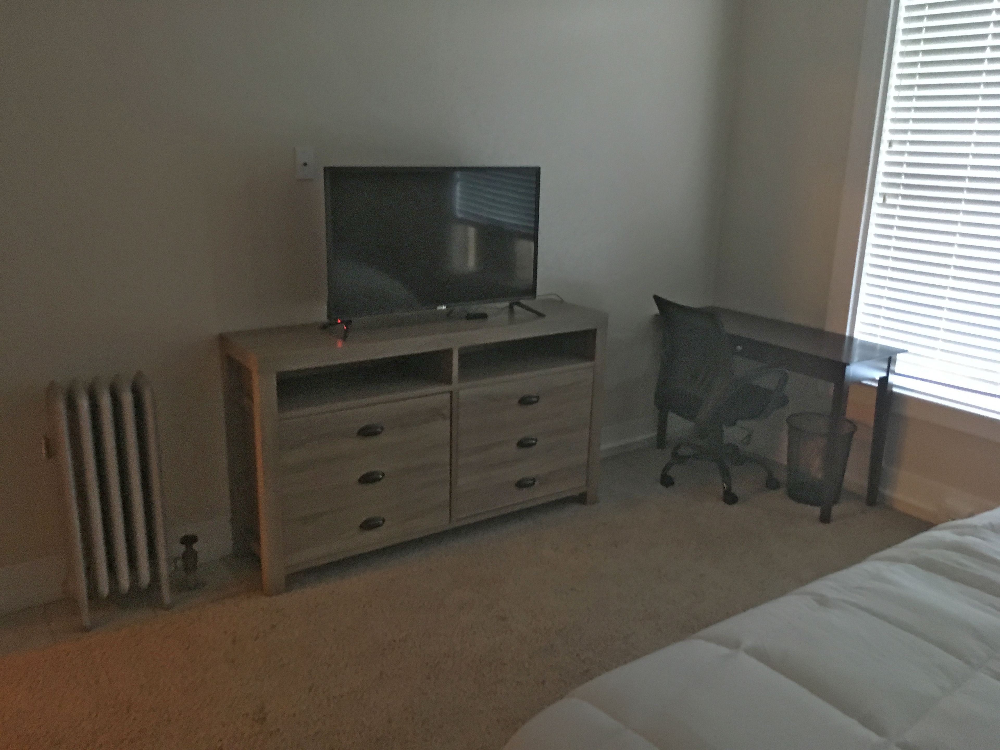 Un dormitorio con cama, cómoda de madera, escritorio y radiador.