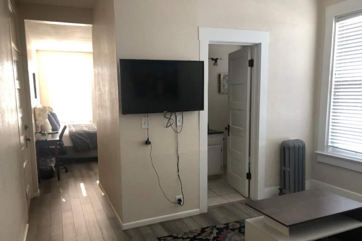 A modern living room with a flat-screen TV mounted on the wall, a wooden floor, and a large window with blinds.