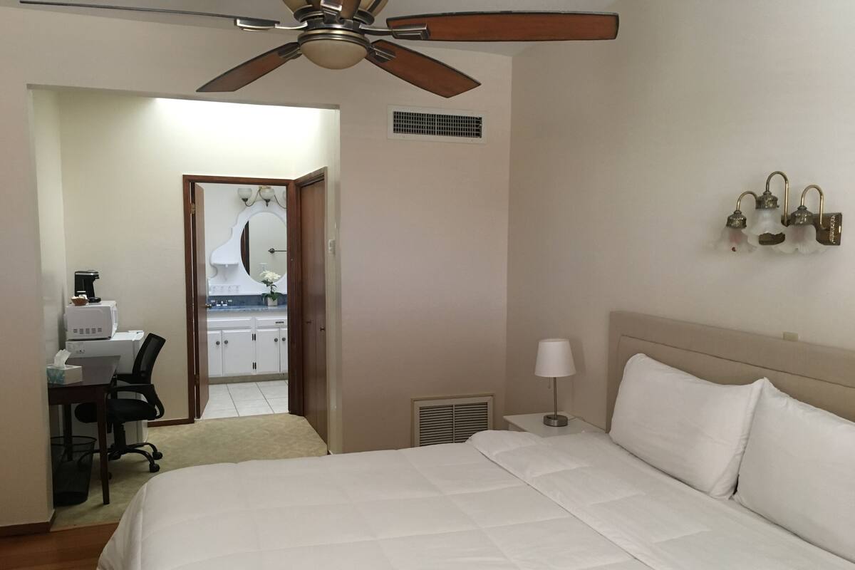 A hotel room with a bed, a desk with a computer, a ceiling fan, and a bathroom visible through an open door.