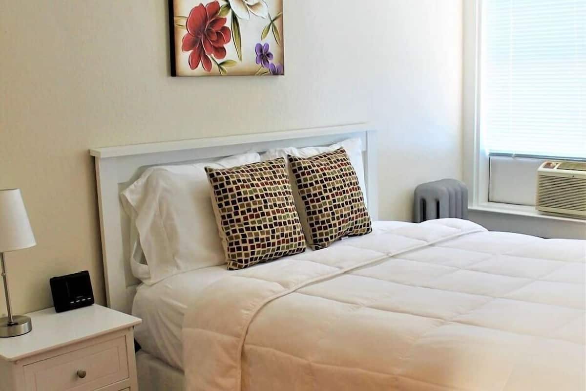 A neatly made bed with a white duvet and two patterned pillows, a bedside table with a lamp and a small electronic device, and a floral painting on the wall.