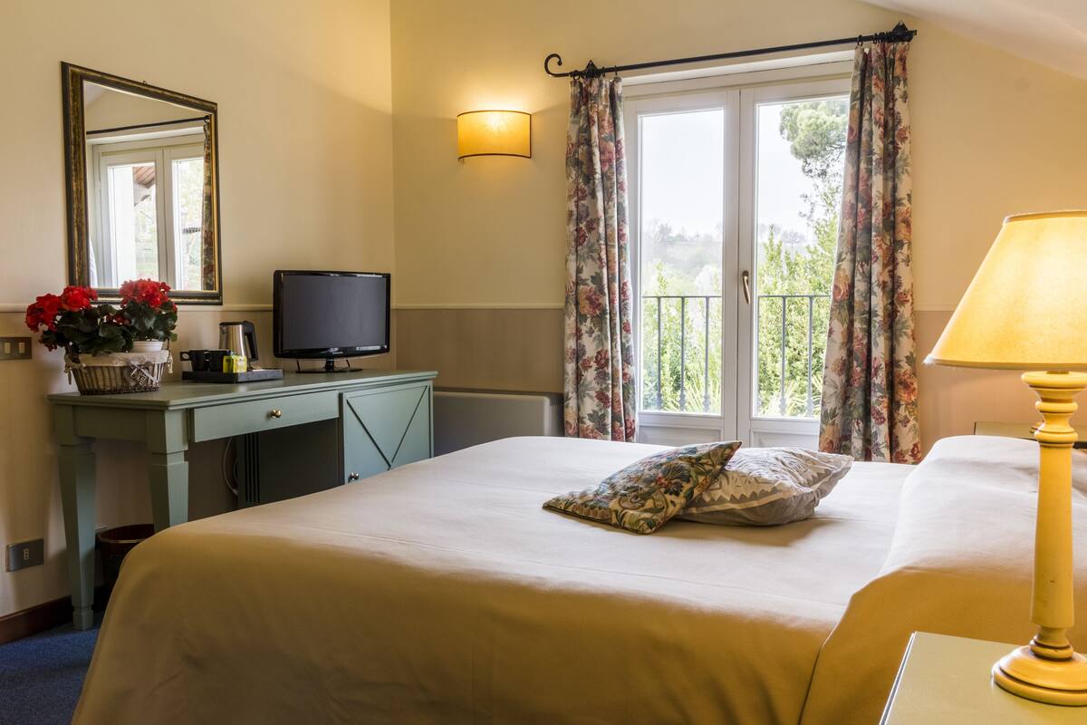 A hotel room with a bed, a desk with a computer, a lamp, a window with curtains, and a view of greenery outside.