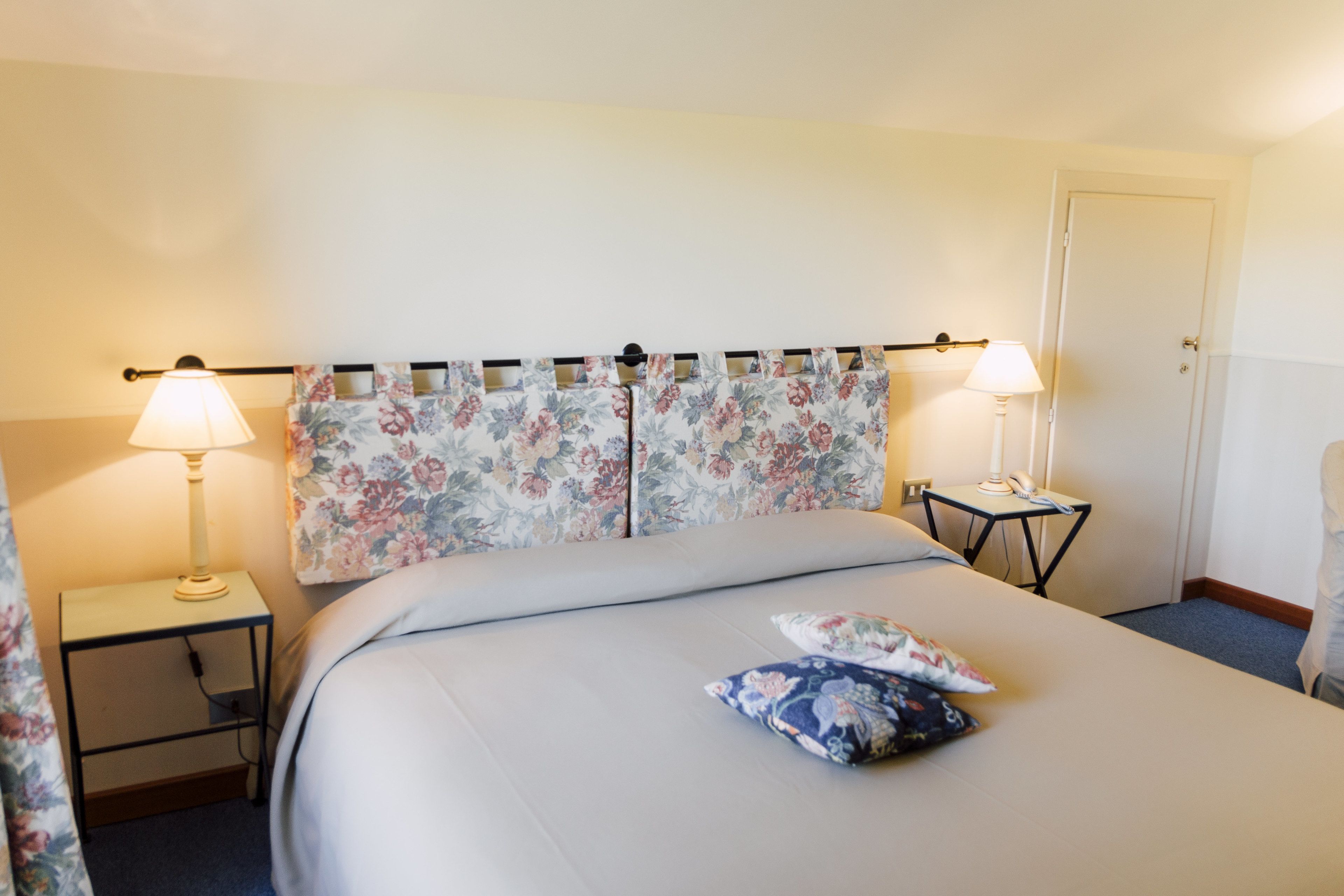 Une chambre d’hôtel avec un lit, deux tables de chevet, une commode, une lampe et une porte.