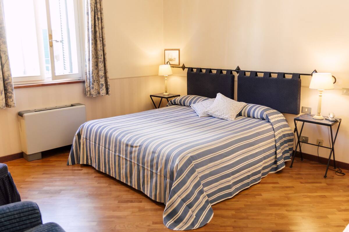 A neatly arranged bedroom with a bed, two bedside tables with lamps, a radiator, and a window with curtains.