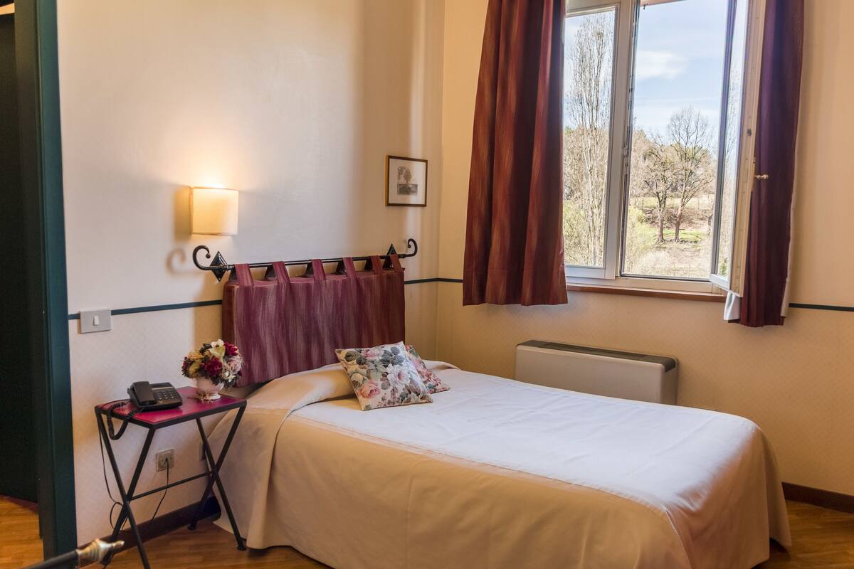 A hotel room with a bed, a small desk with a telephone and a vase of flowers, a radiator, and a window with curtains.
