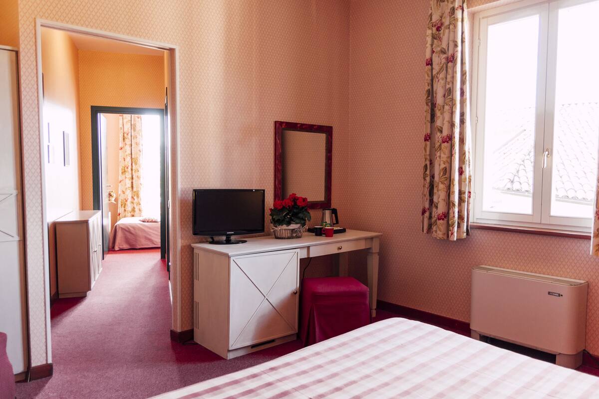 A hotel room with a bed, a desk with a computer, a radiator, and a window with curtains.