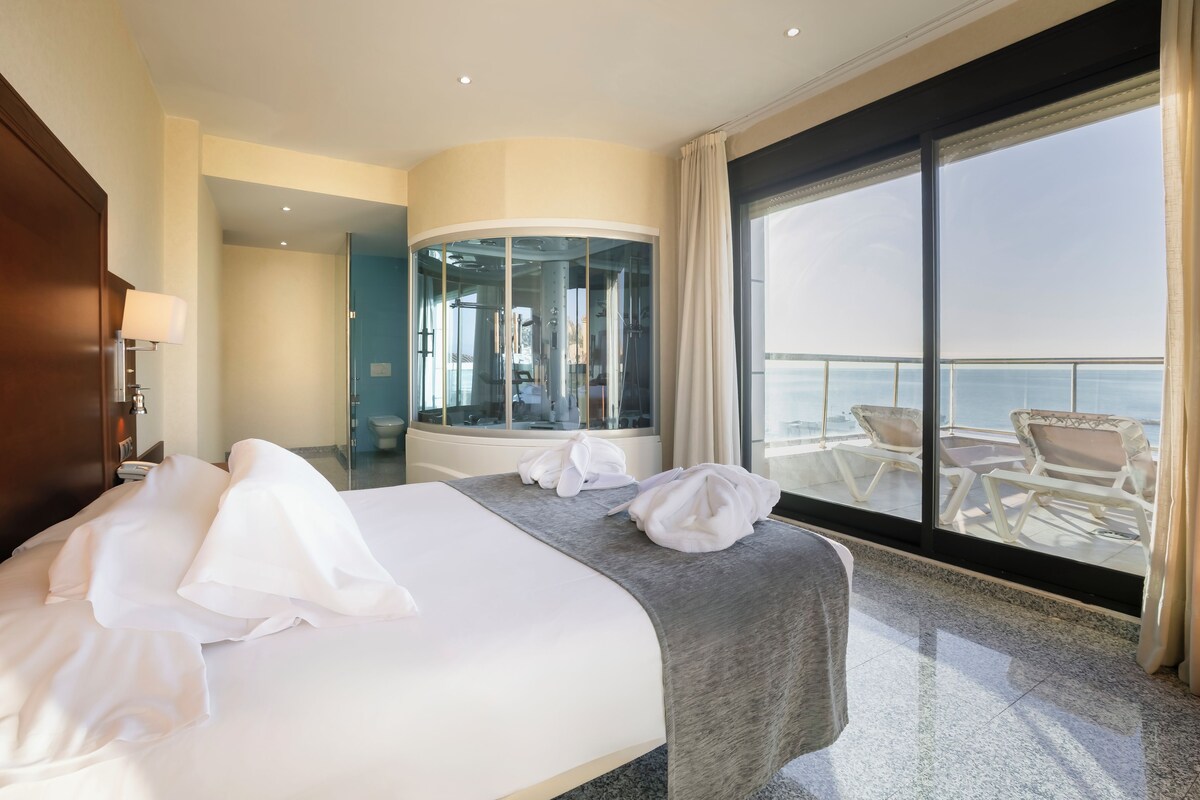 Habitación de hotel con cama, baño con paredes de cristal y balcón con sillas.