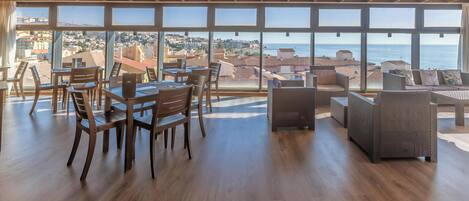Amplia habitación con vigas de madera en el techo, grandes ventanales con vistas al mar y a la ciudad, y mobiliario de mesas y sillas de madera.
