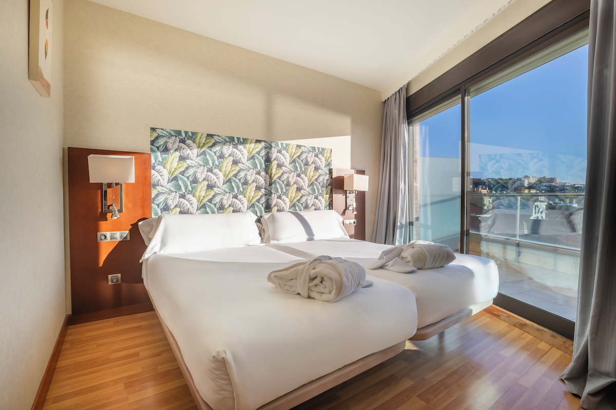 Habitación de hotel con dos camas, un ventanal con vistas y un cuadro de temática tropical.