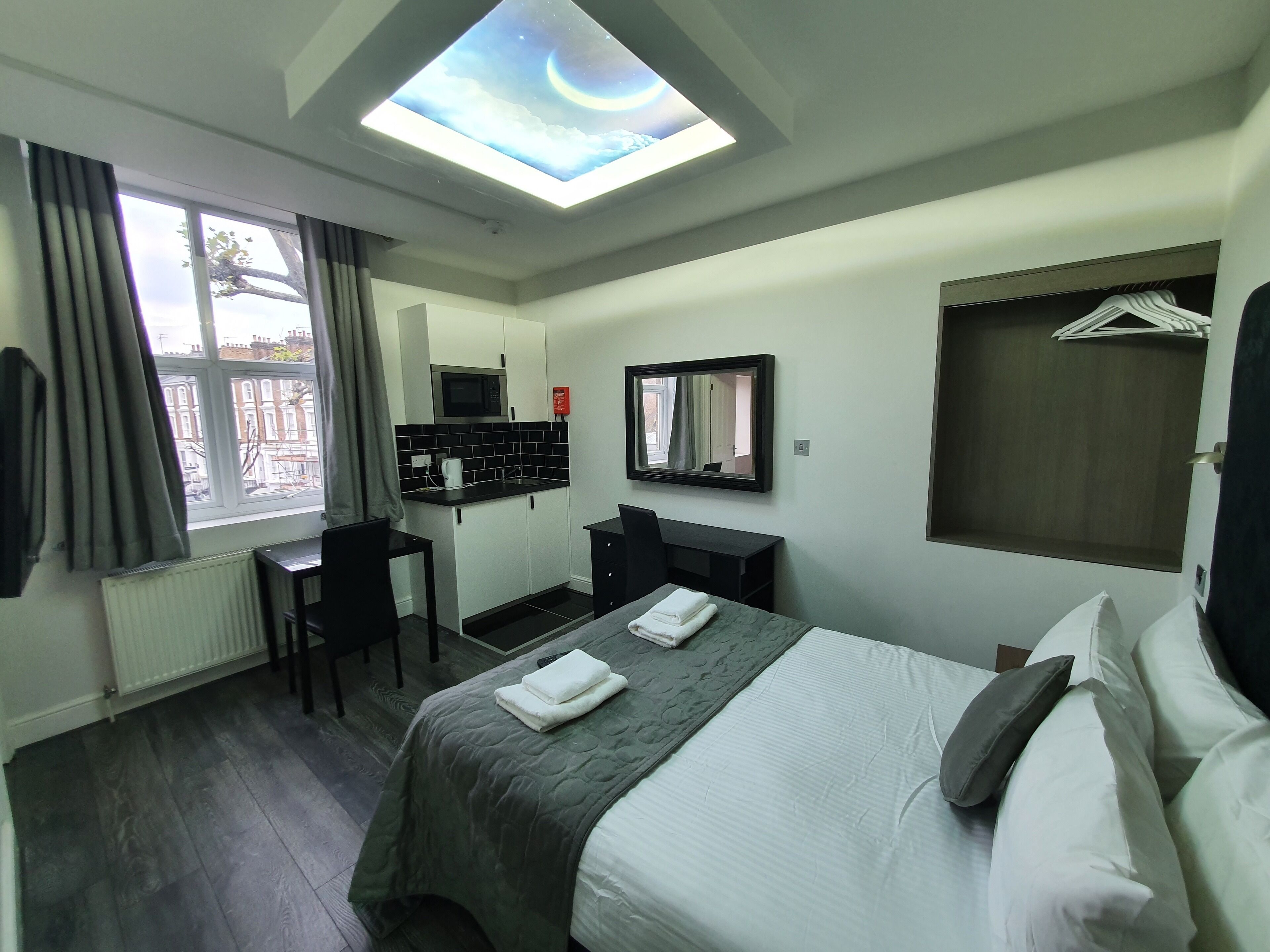 A hotel room with a skylight, a bed with pillows, a desk with a chair, a small kitchenette, and a mirror.