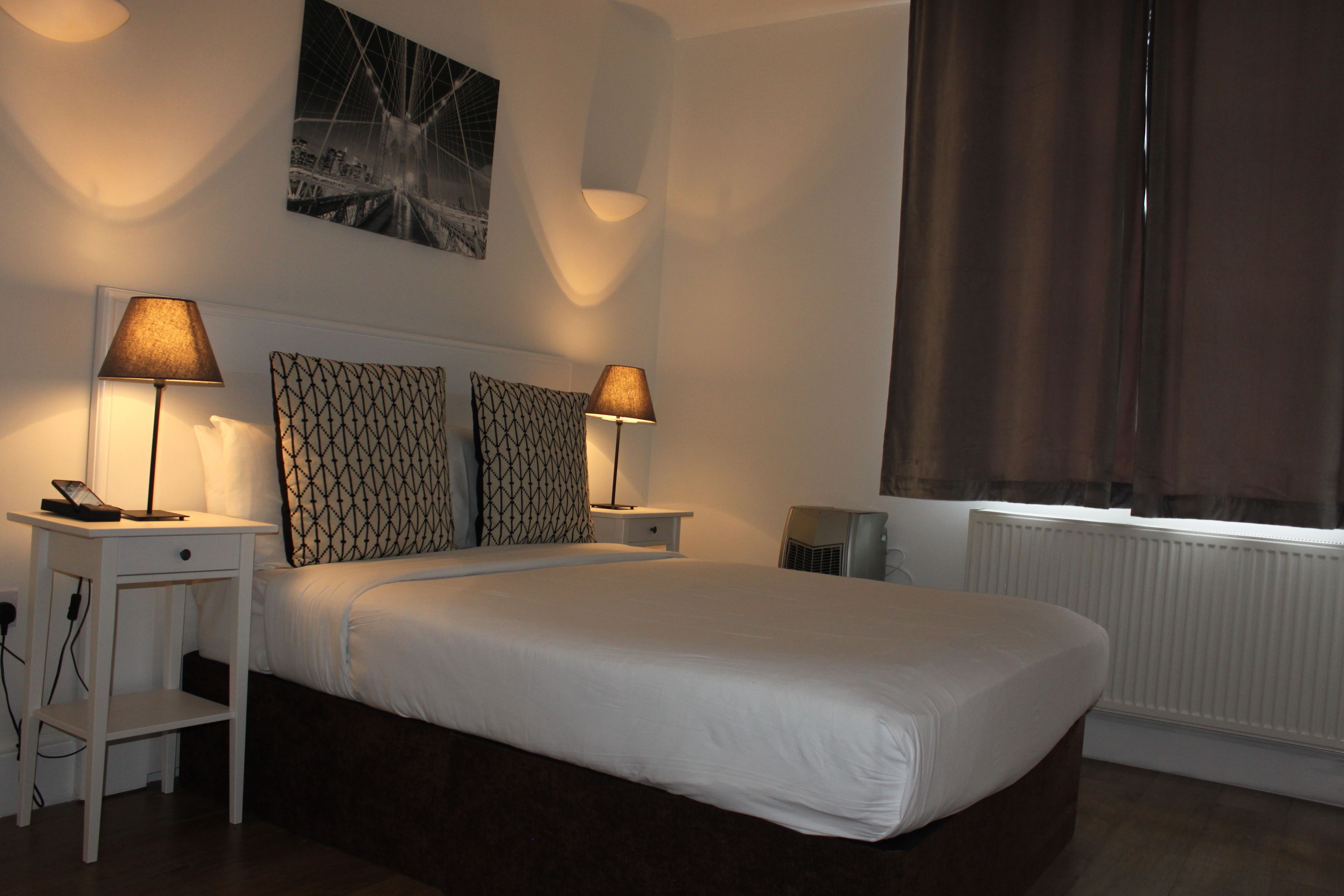 A neatly made bed with white linens, two bedside lamps, a patterned headboard, a small bedside table with a lamp, and a window with curtains.
