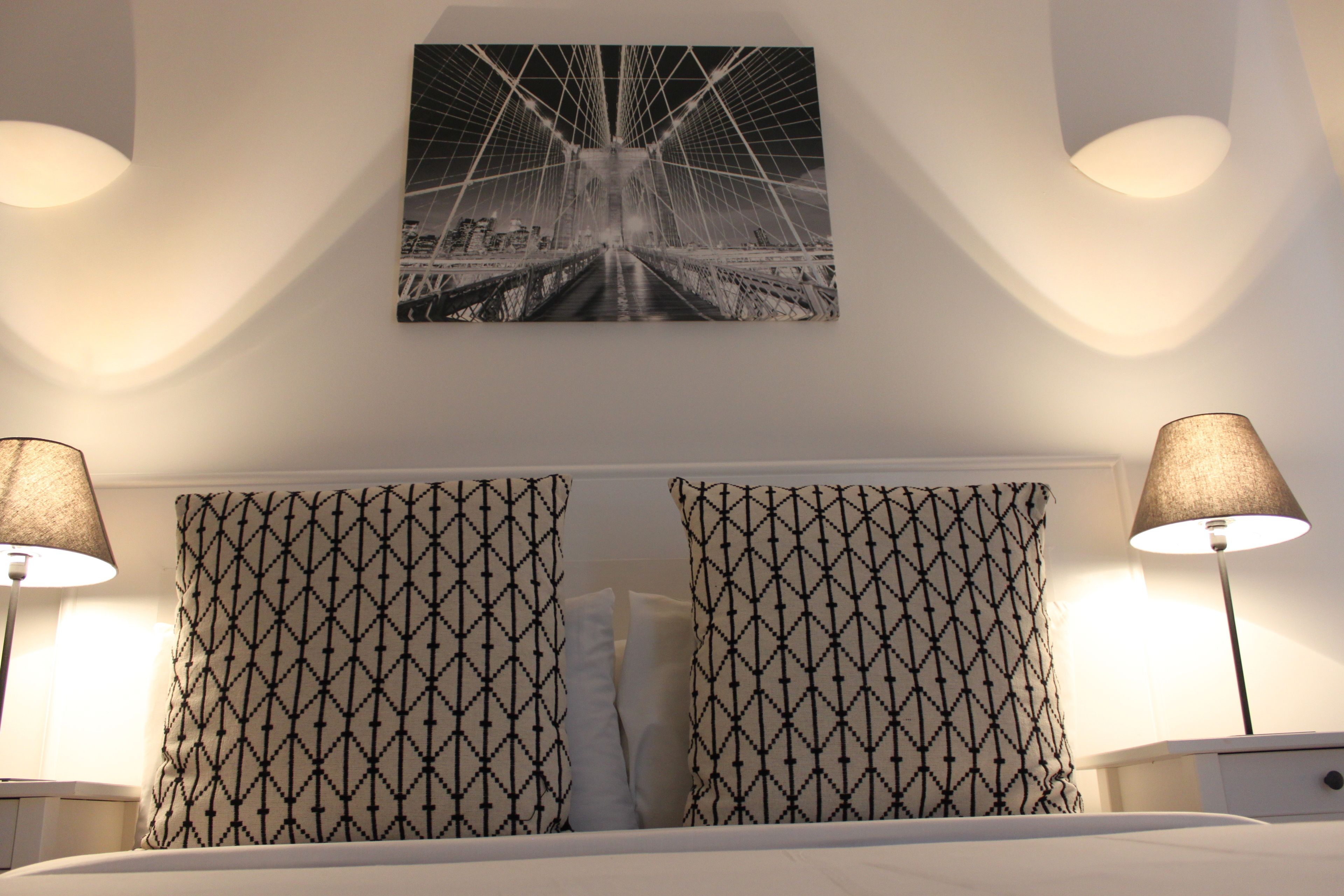 Habitación de hotel con una cama, dos almohadones estampados, un cuadro colgado en la pared de un puente y dos lámparas de noche.
