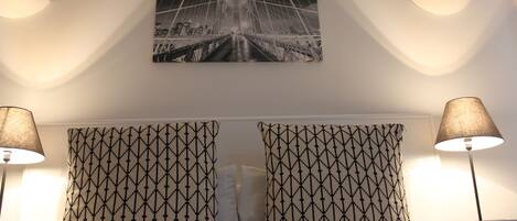 Habitación de hotel con una cama, dos almohadones estampados, un cuadro colgado en la pared de un puente y dos lámparas de noche.