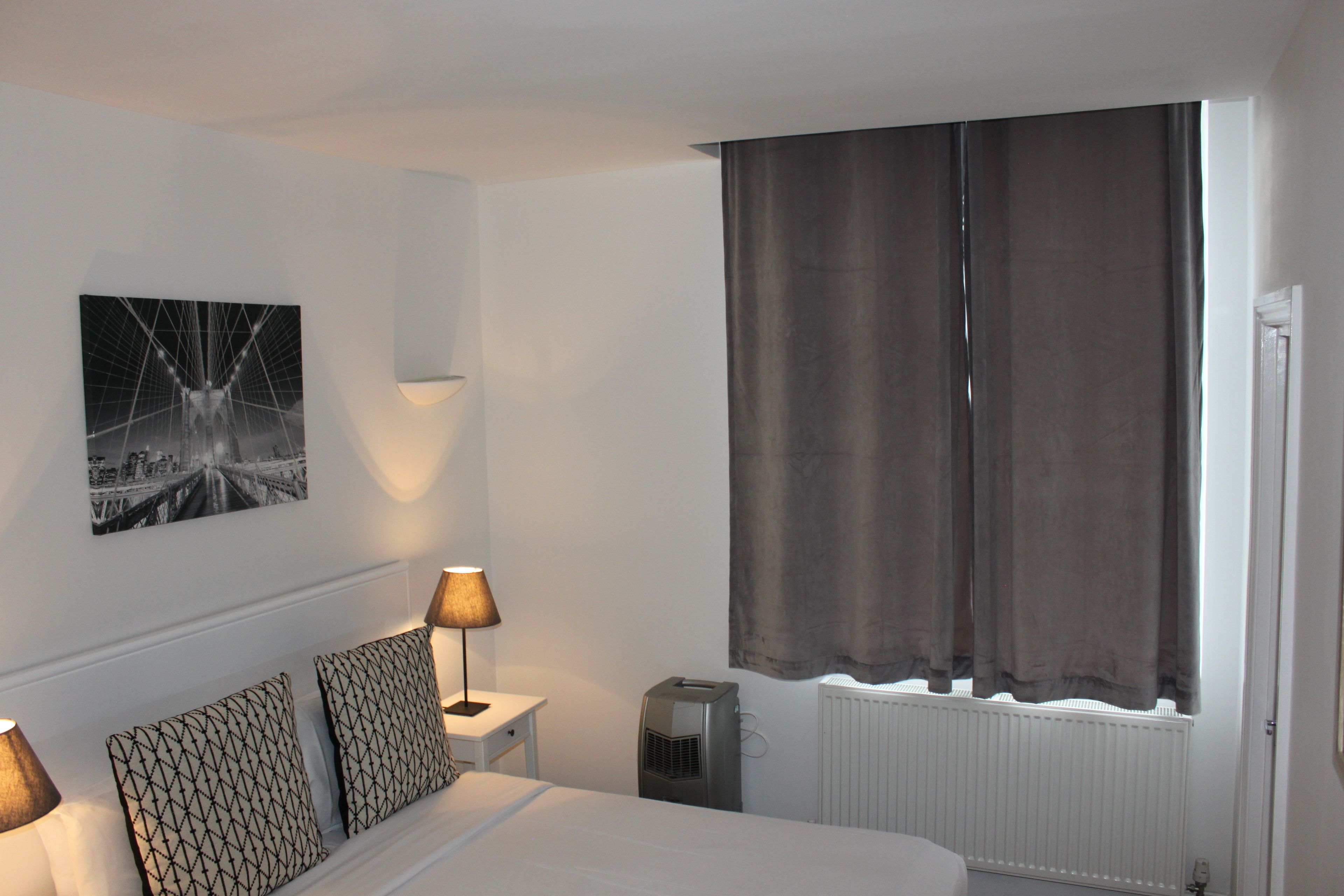 A hotel room with a bed, two bedside lamps, a wall-mounted picture, a radiator, and a window with curtains.