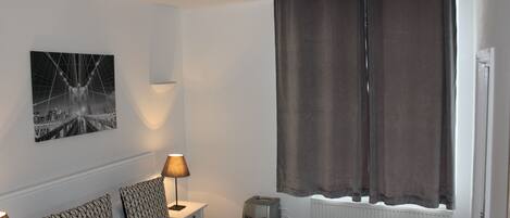 A hotel room with a bed, two bedside lamps, a wall-mounted picture, a radiator, and a window with curtains.