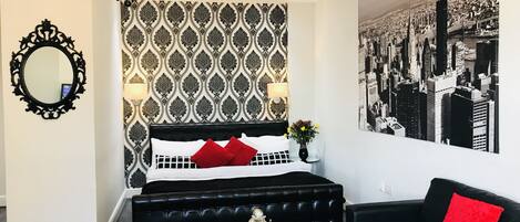 A modern bedroom with a patterned wallpaper, a black upholstered bed, a red pillow, a cityscape mural, a chandelier, and a round mirror.