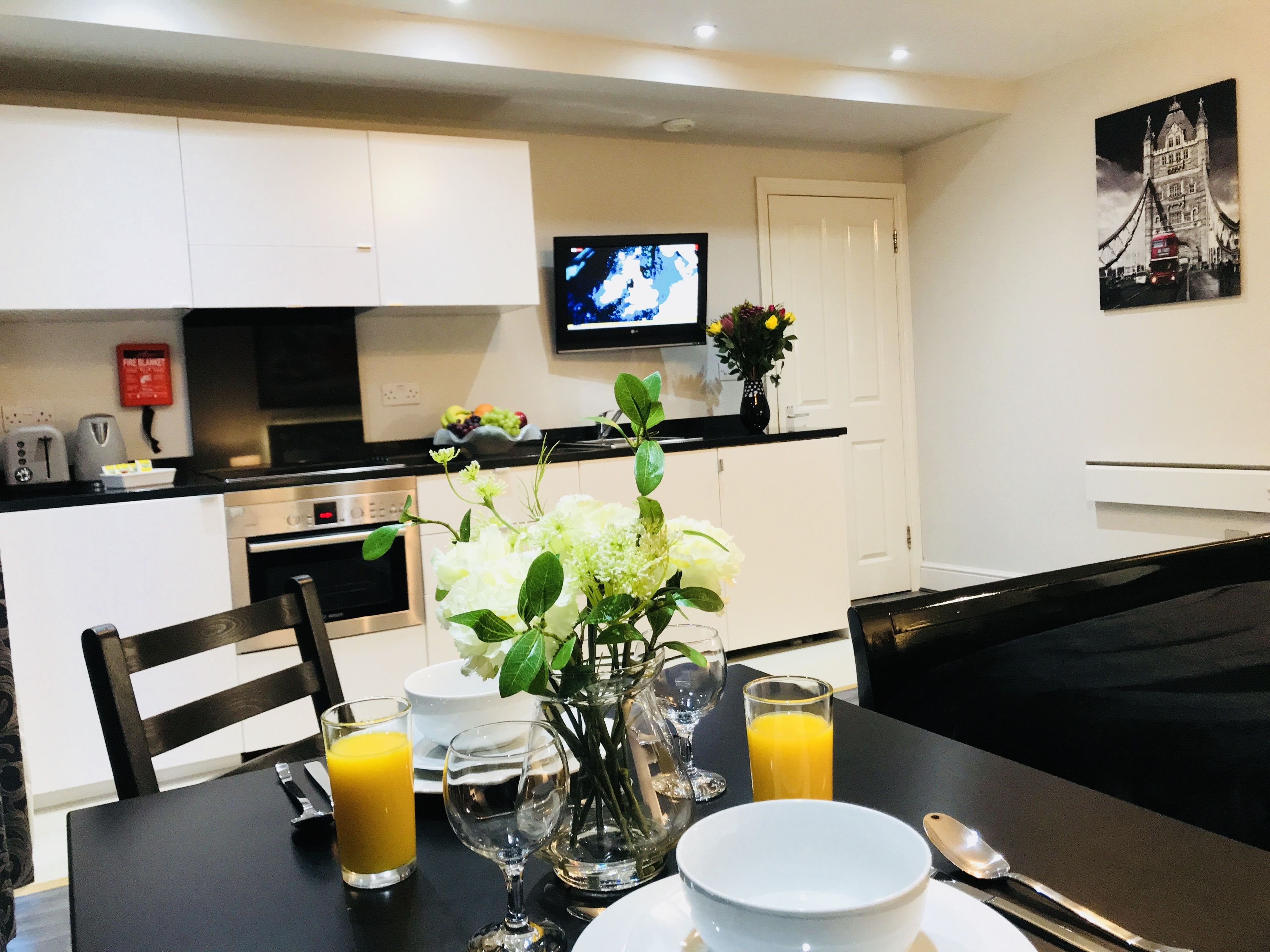 A dining area with a table set for two, a vase with flowers, and a television mounted on the wall.