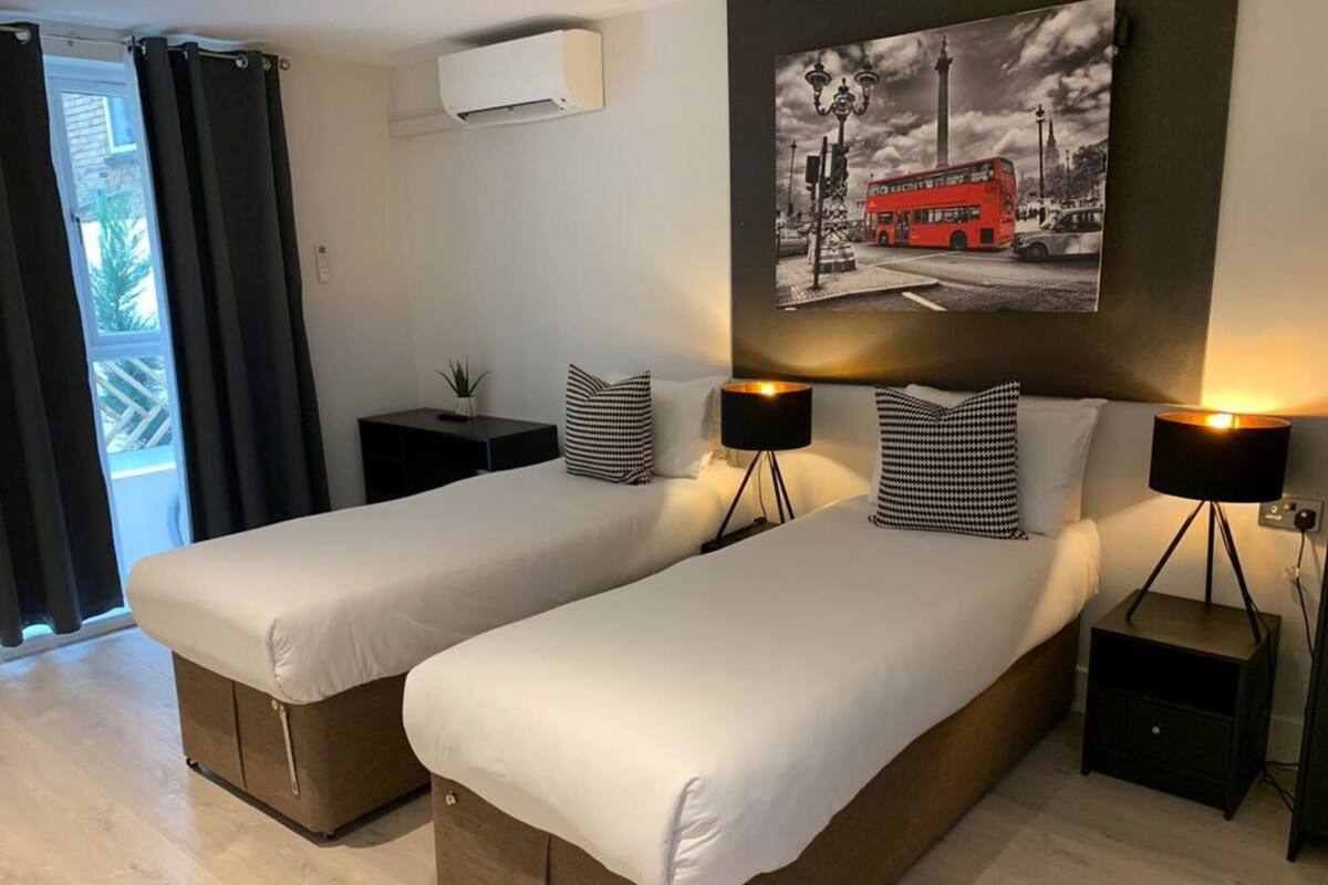 A hotel room with two beds, a small bedside table with a lamp, a wall-mounted air conditioner, and a large window with curtains.
