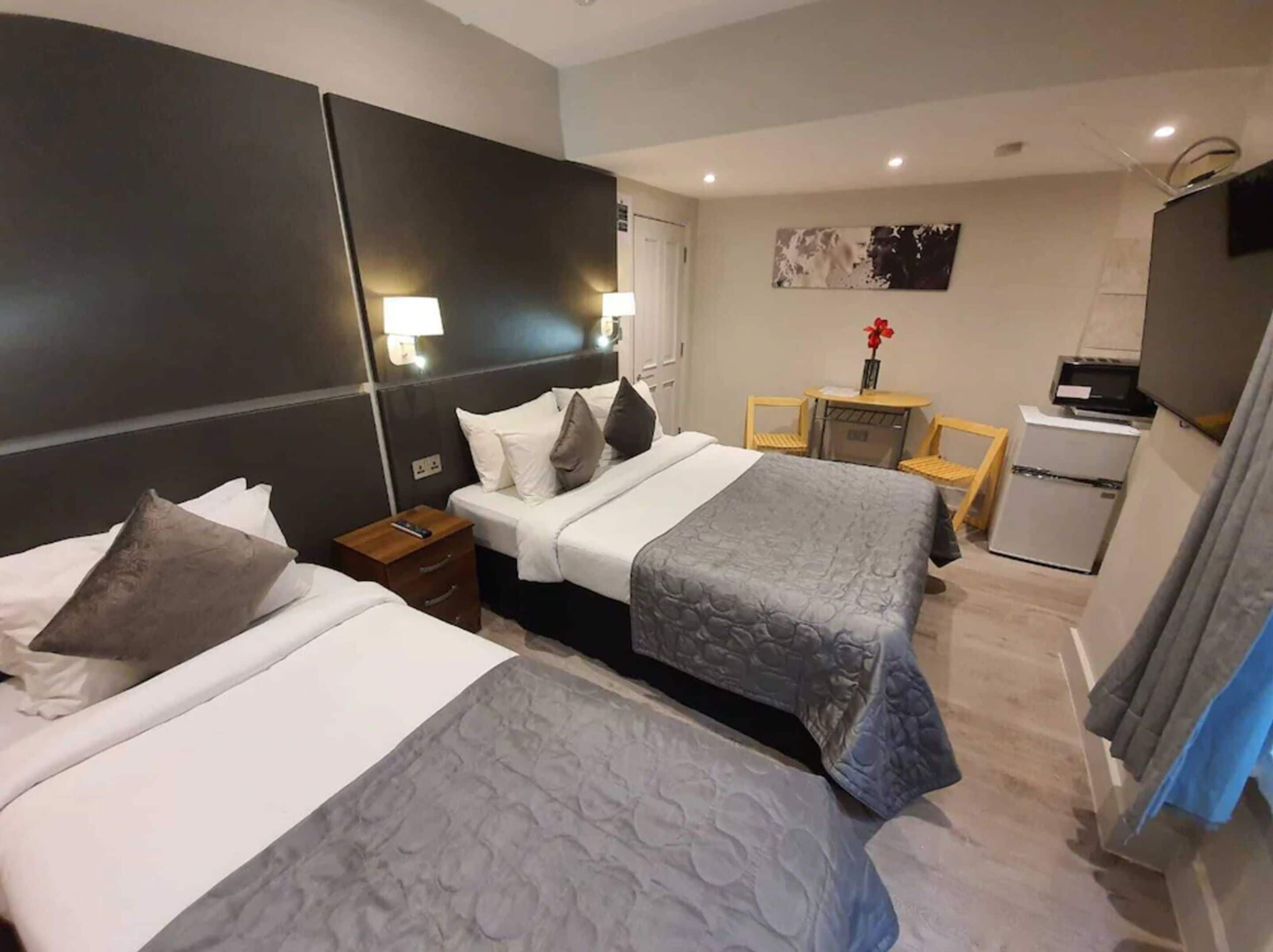 Ein Hotelzimmer mit zwei Betten, einem kleinen Tisch, einem Sessel, einem Schreibtisch, einem Kühlschrank und einem Fernseher.