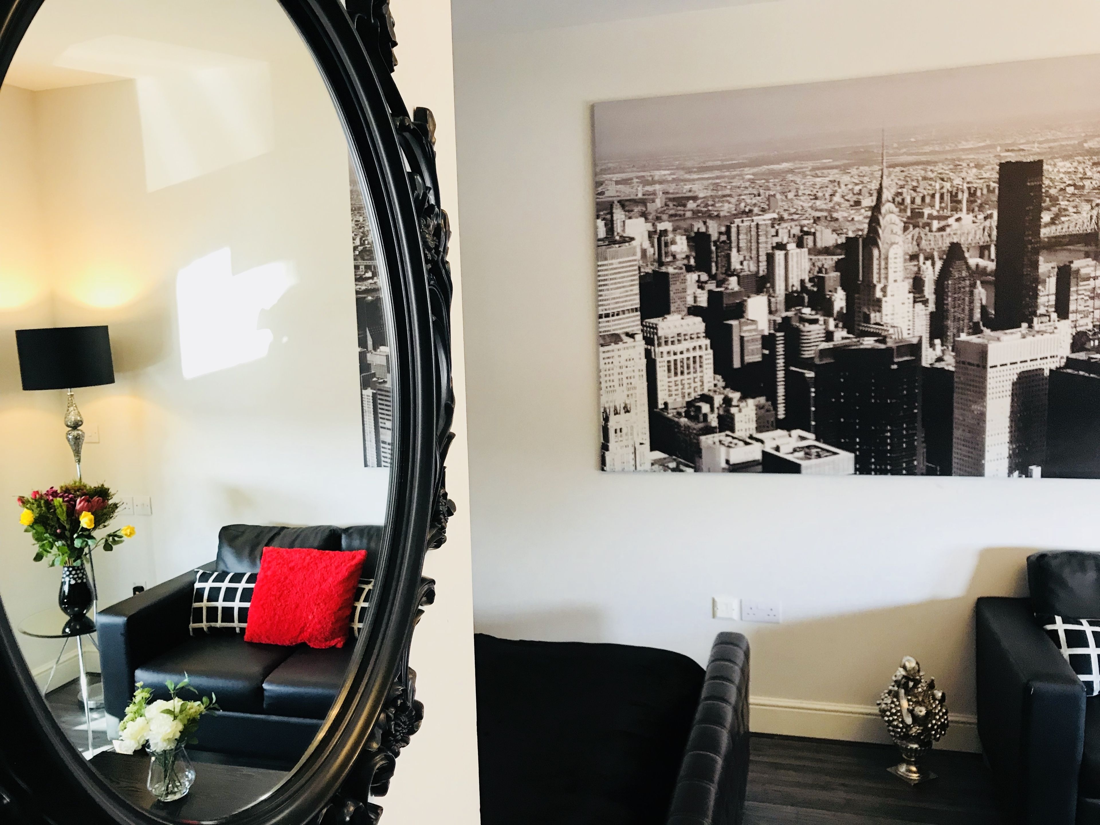 A living room with a black sofa, a red pillow, a black-framed mirror, and a black-and-white cityscape painting on the wall.