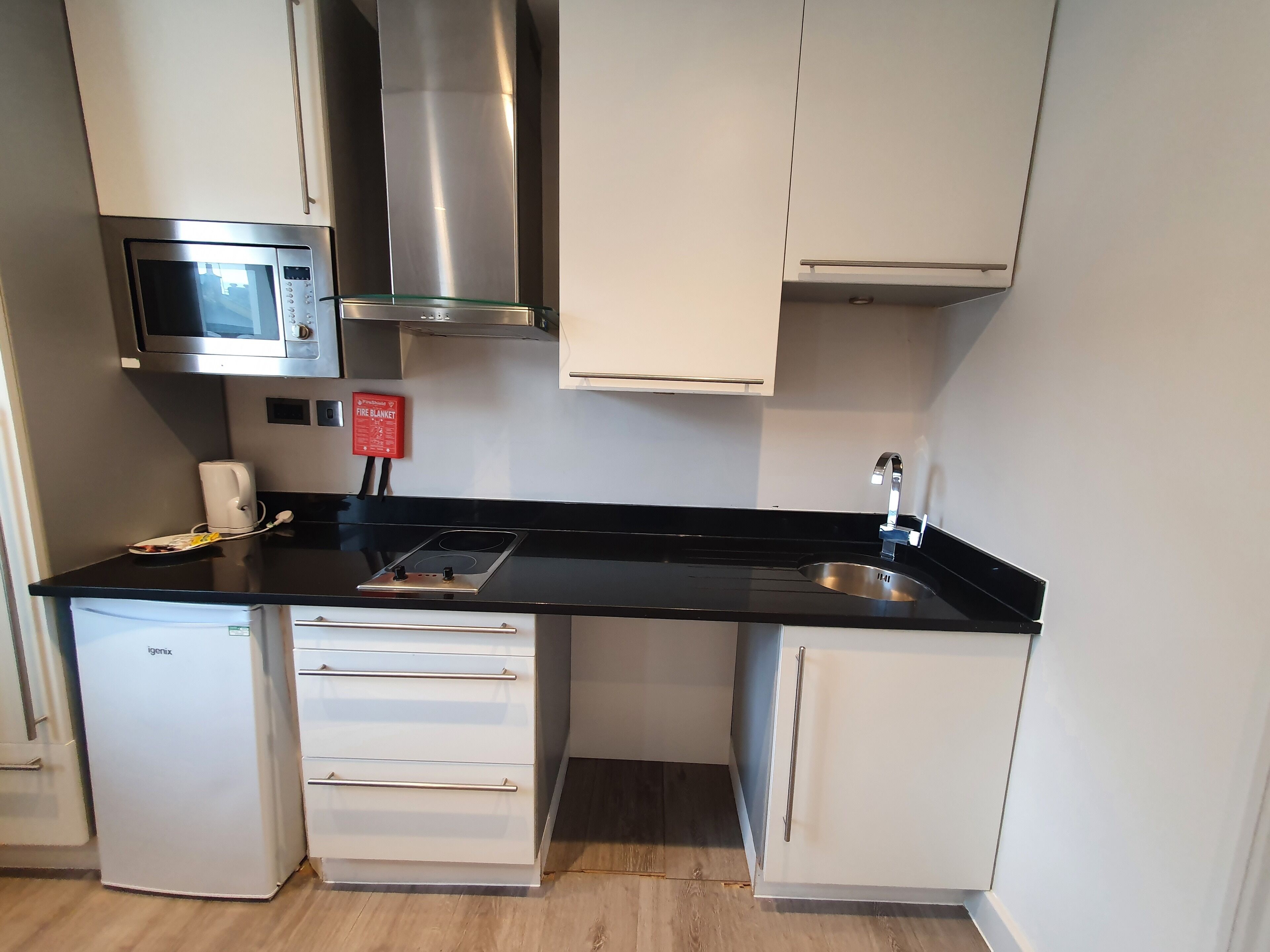 A compact kitchen with a microwave, gas stove, and a sink. There is a fire blanket on the wall and a small refrigerator.