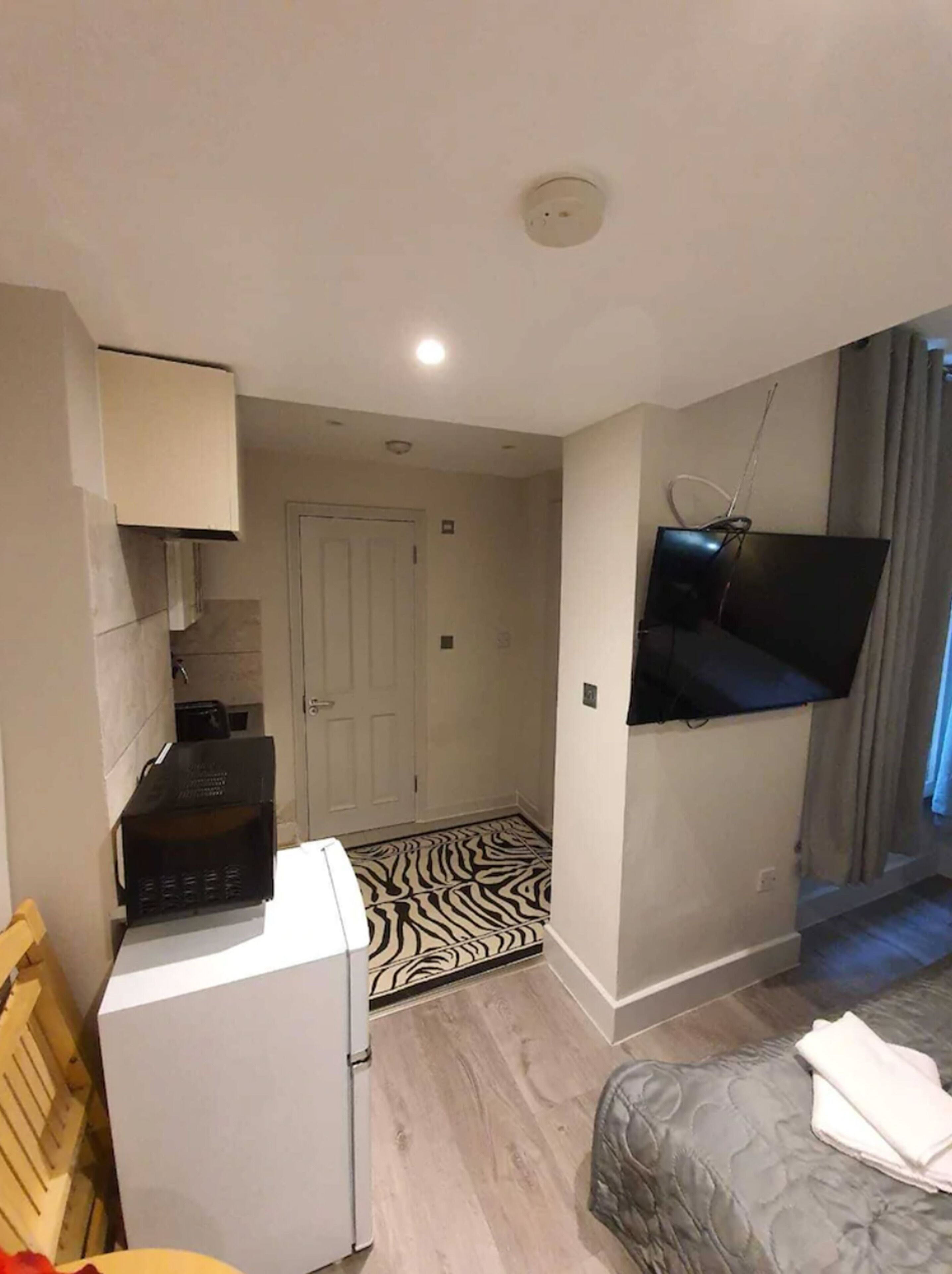 A compact hotel room with a bed, a TV mounted on the wall, a small kitchenette, and a zebra-patterned rug.