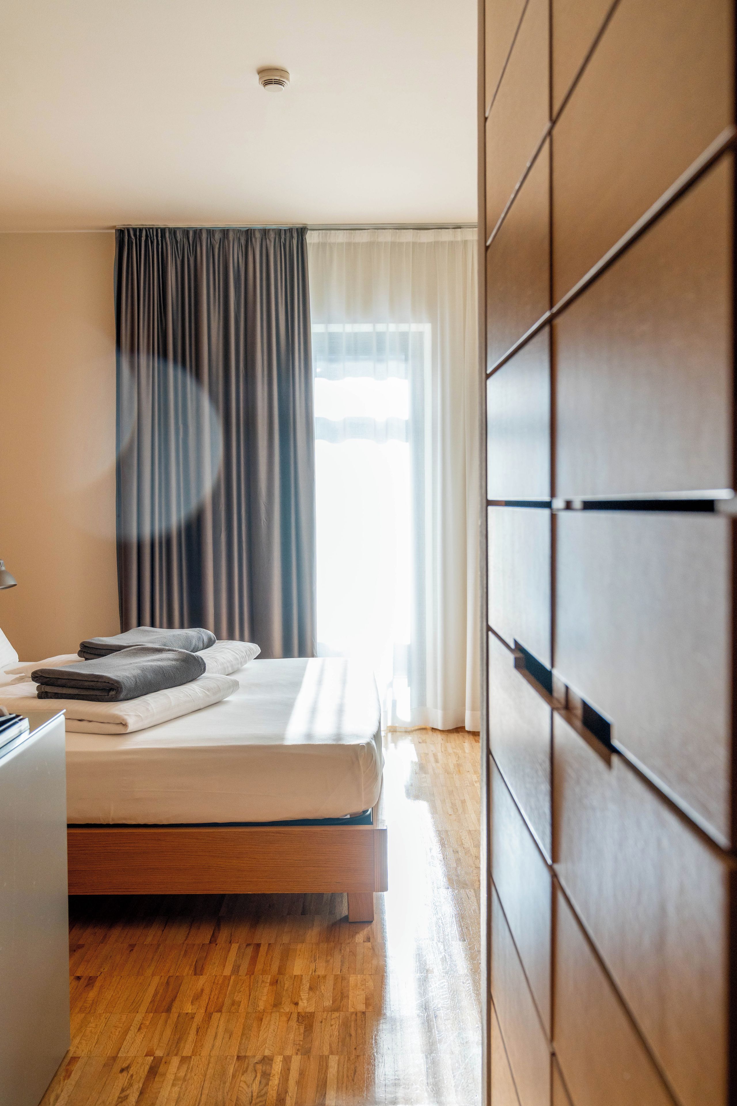 Een moderne slaapkamer met een houten vloer, een bed met een wit matras en grijze kussens, een nachtkastje en een raam met gordijnen.