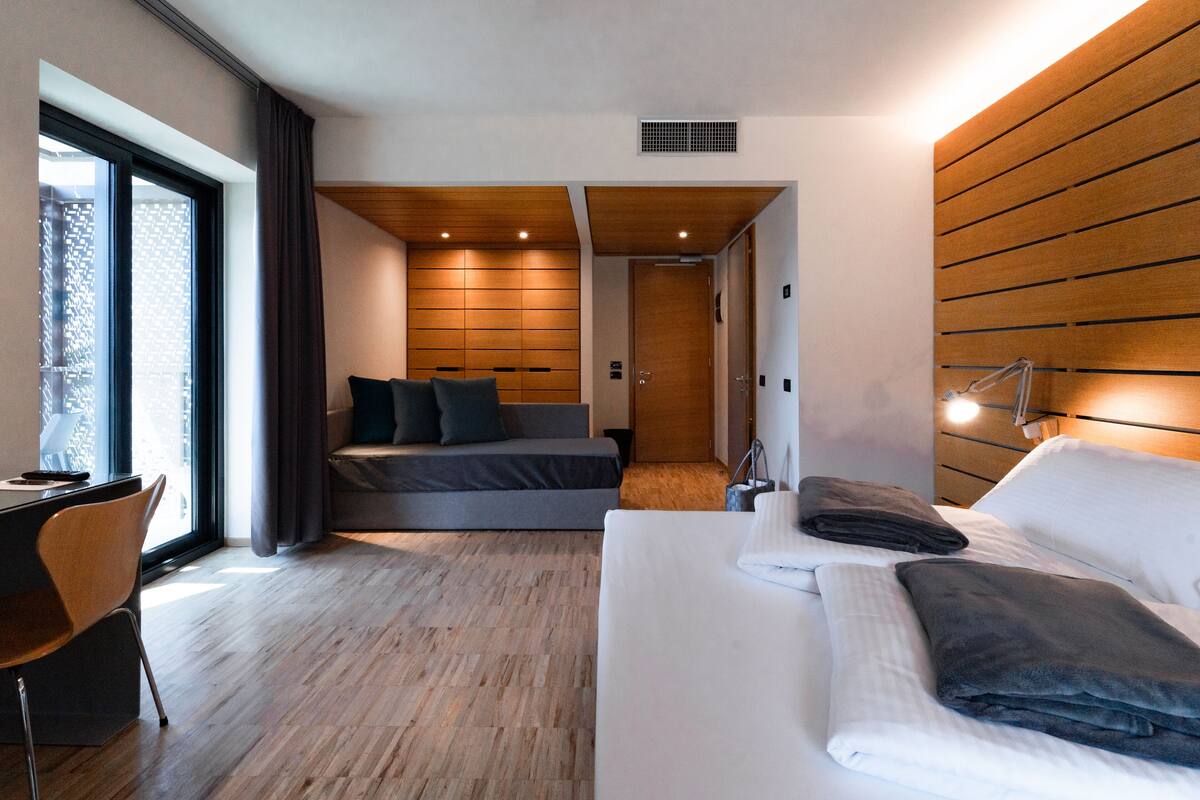 A modern hotel room with a bed, a sofa, a desk, and a large window.