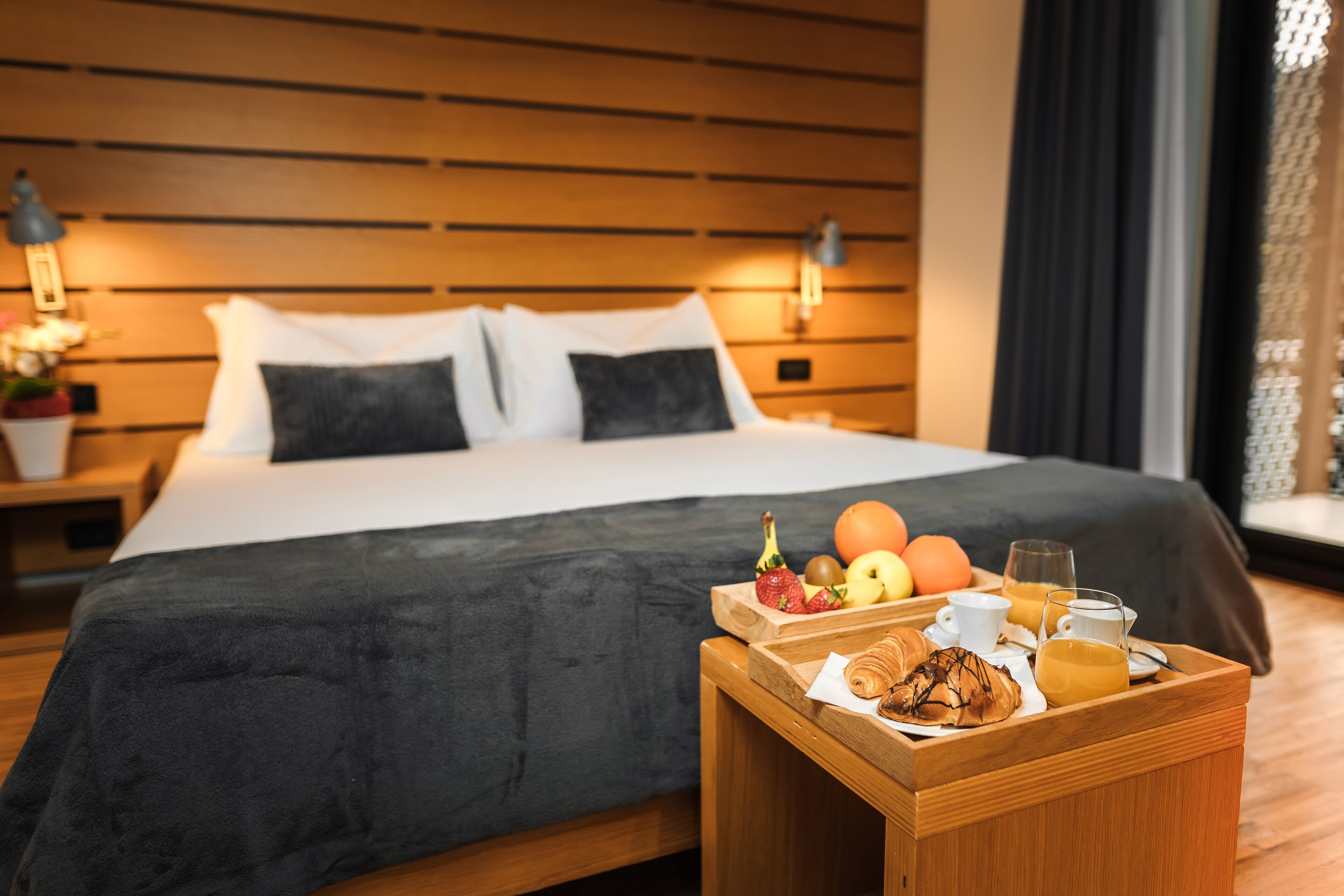 Una cama bien hecha con una colcha oscura y dos almohadas, una mesita de noche de madera con una bandeja de desayuno, y una pared con paneles de madera horizontales.
