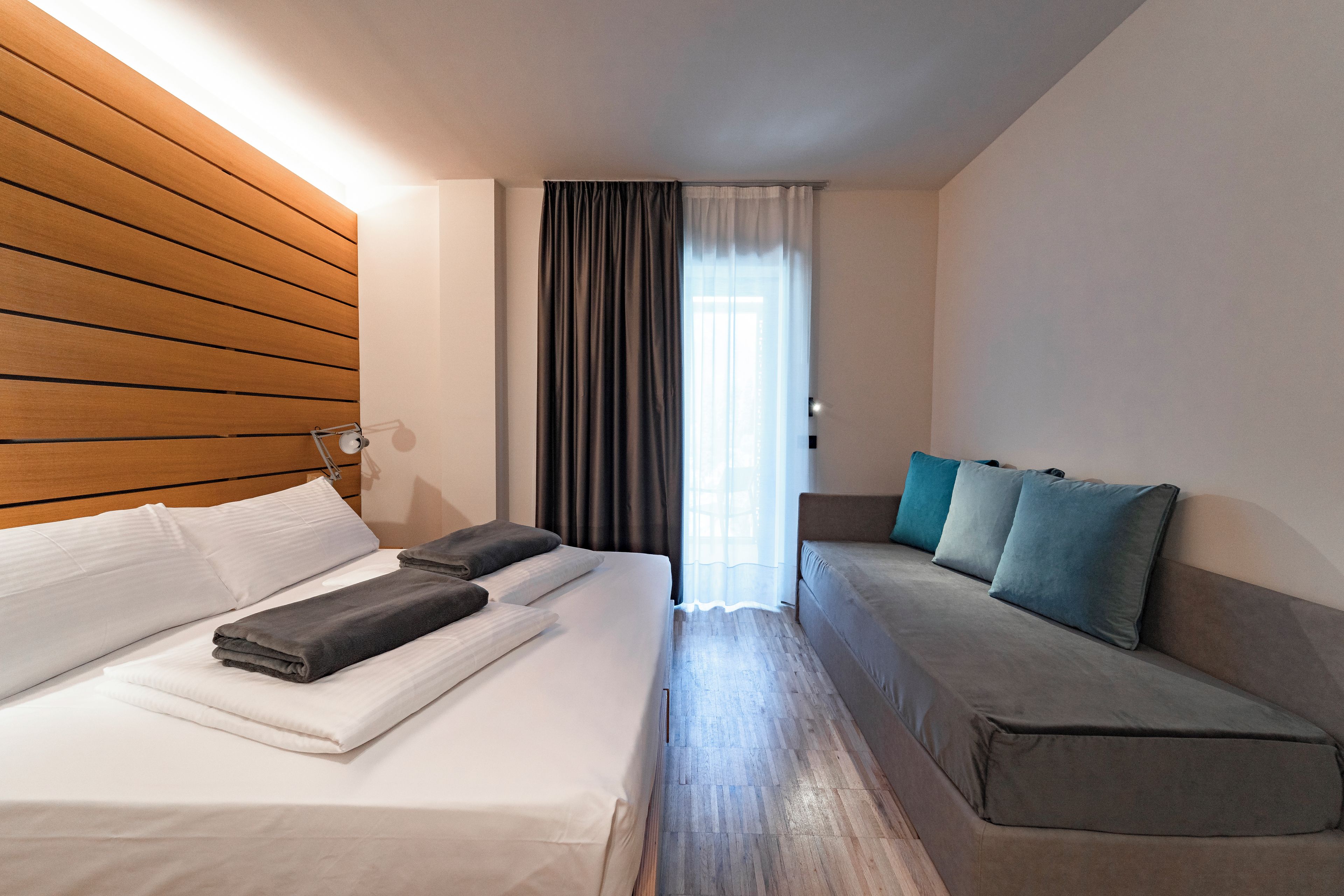 Ein Hotelzimmer mit Bett, Nachttisch, Lampe, einem Fenster mit Vorhängen und einer Couch mit Kissen.