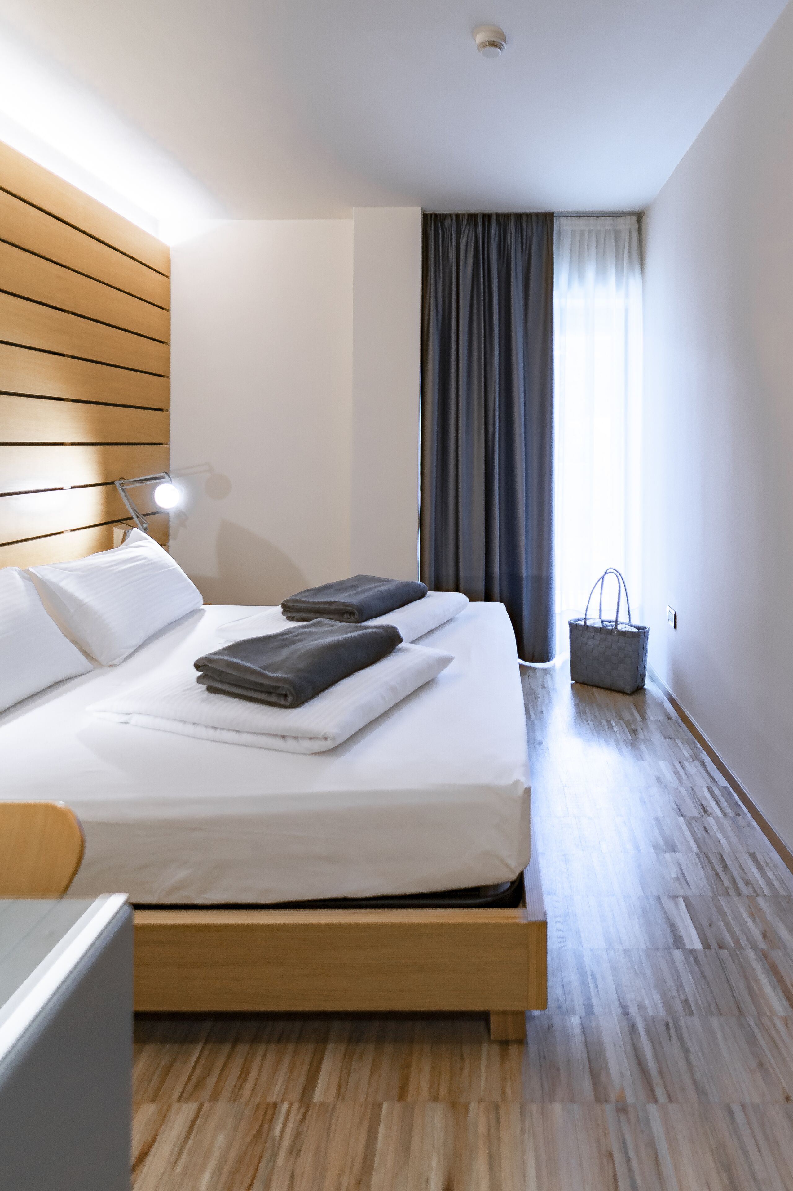 Una cama bien hecha con almohadas y toallas dobladas, una lámpara de noche y una bolsa gris en el suelo.