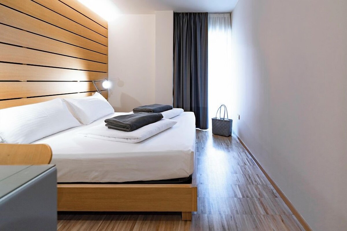 A modern hotel room with a wooden headboard, a neatly made bed, a bedside table with a lamp, and a view of the hallway.