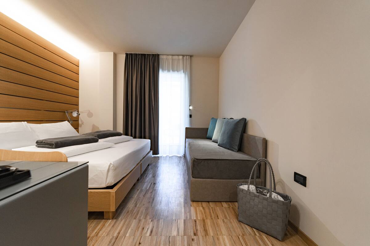 A modern hotel room with a wooden headboard, a bed with white linens, a grey sofa, a small desk, and a grey handbag on the floor.