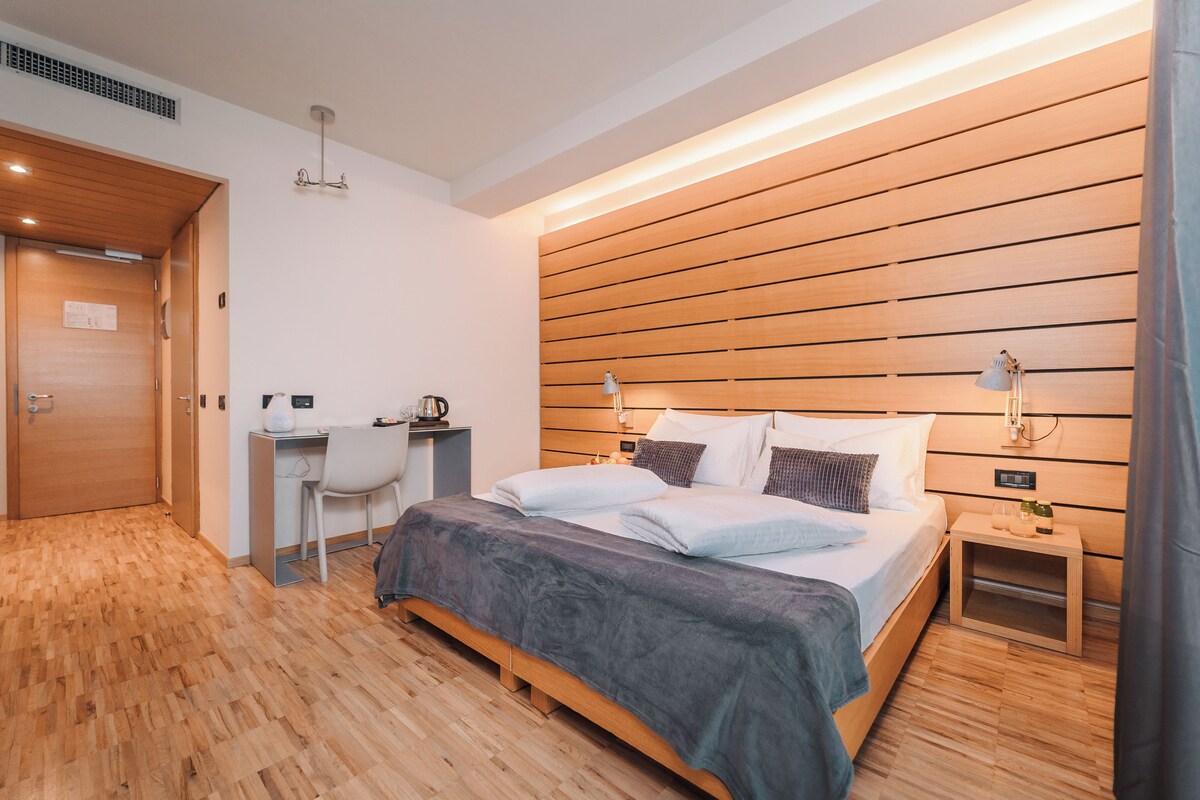 A modern hotel room with a wooden floor, a desk with a chair, a bed with a grey blanket, and a nightstand with a lamp.