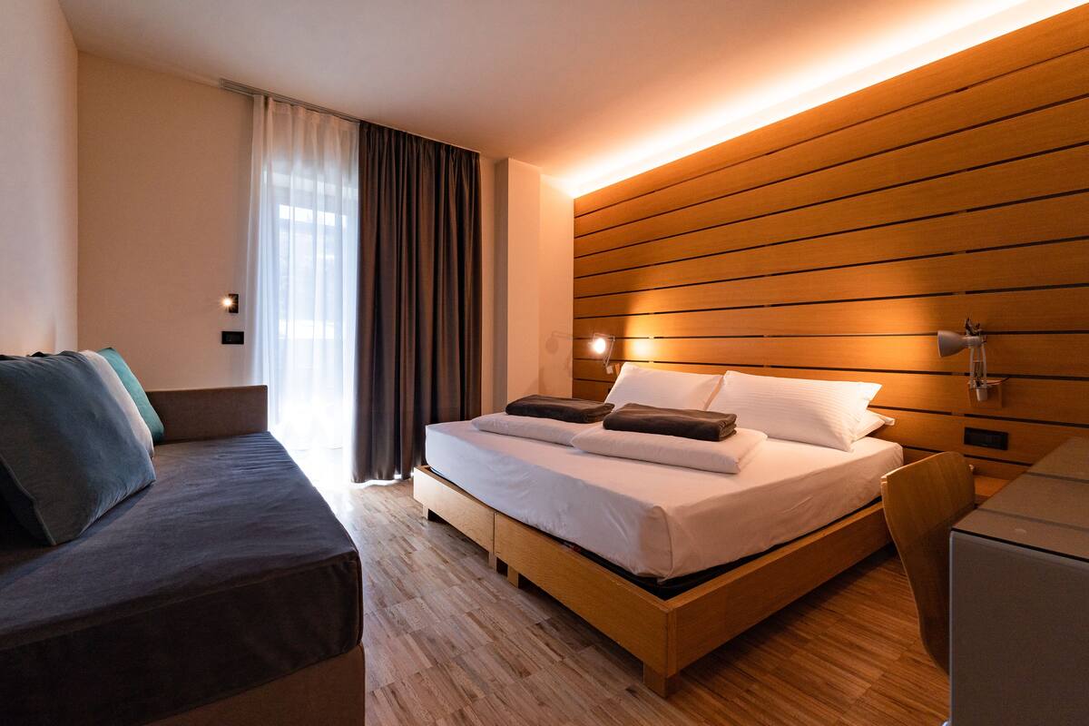 A modern hotel room with a wooden bed, a wooden nightstand, a desk, and a sofa.