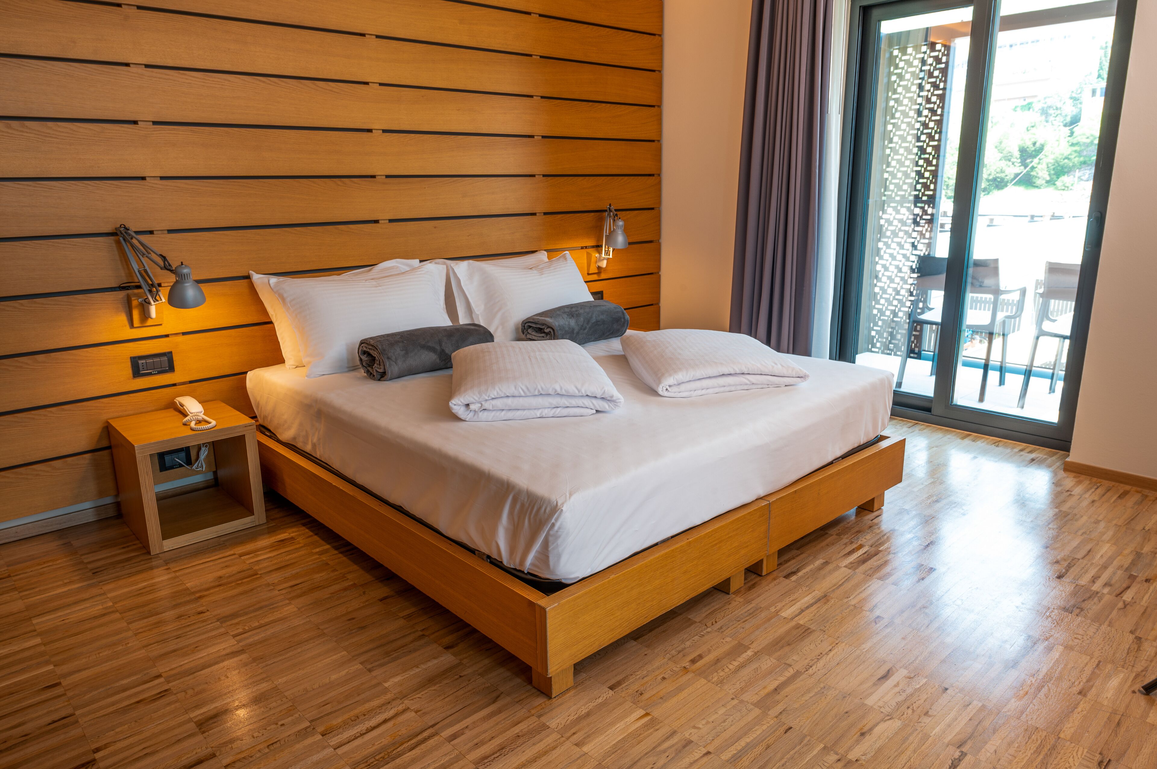A modern bedroom with a wooden headboard, a wooden bed frame, a nightstand with a phone, and a view of the outdoors through a glass door.
