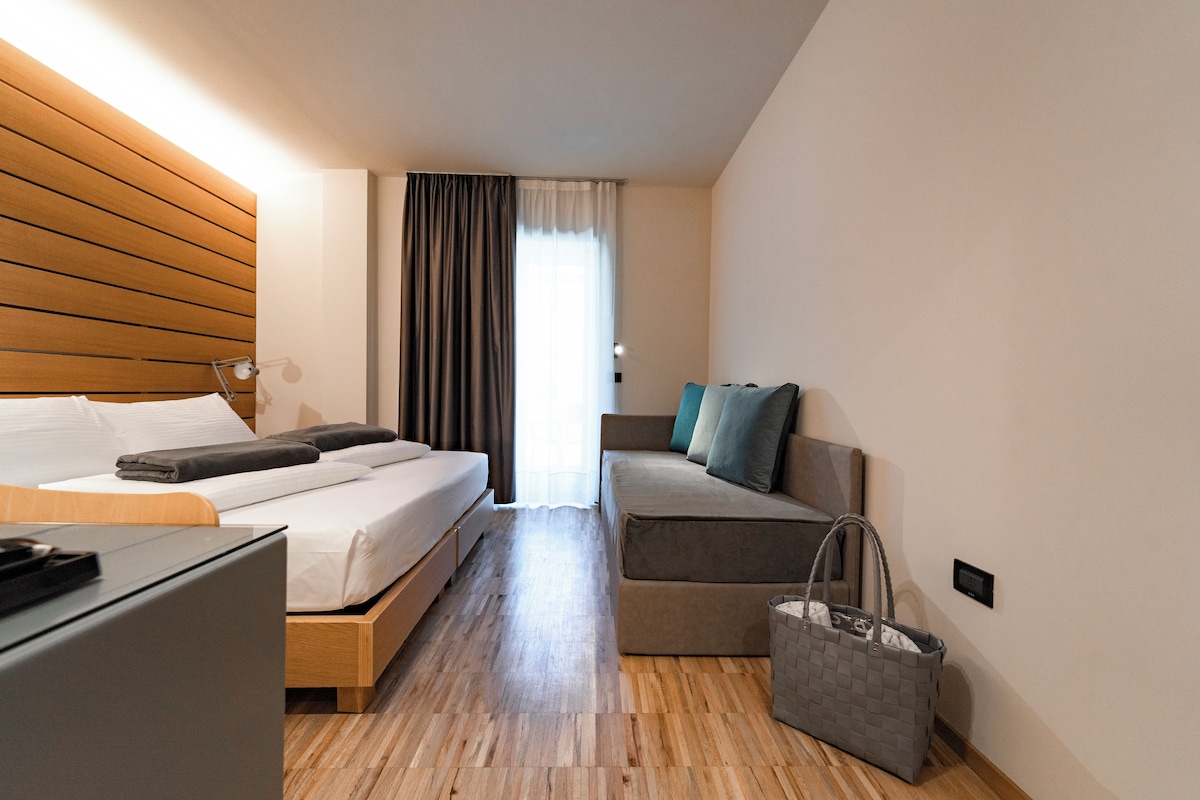 Una habitación de hotel moderna con cabecera de madera, cama con ropa de cama blanca, sofá gris, un escritorio pequeño y un bolso gris en el suelo.