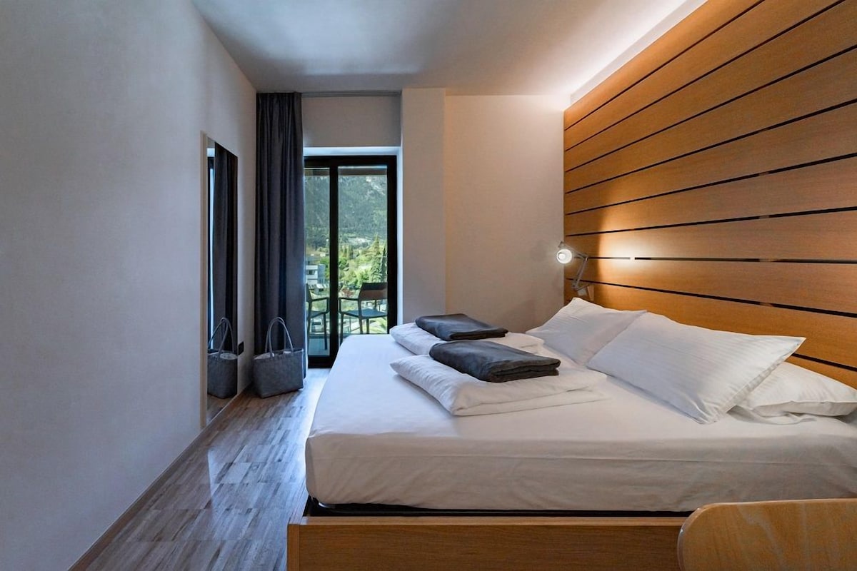 Un dormitorio moderno con cabecera de madera, un ventanal con cortinas y una cama bien hecha con almohadas y un edredón doblado.