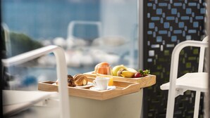 En bakke med morgenmad, blandt andet croissanter, frugt og en kop kaffe, placeret på et bord med udsigt til havet.