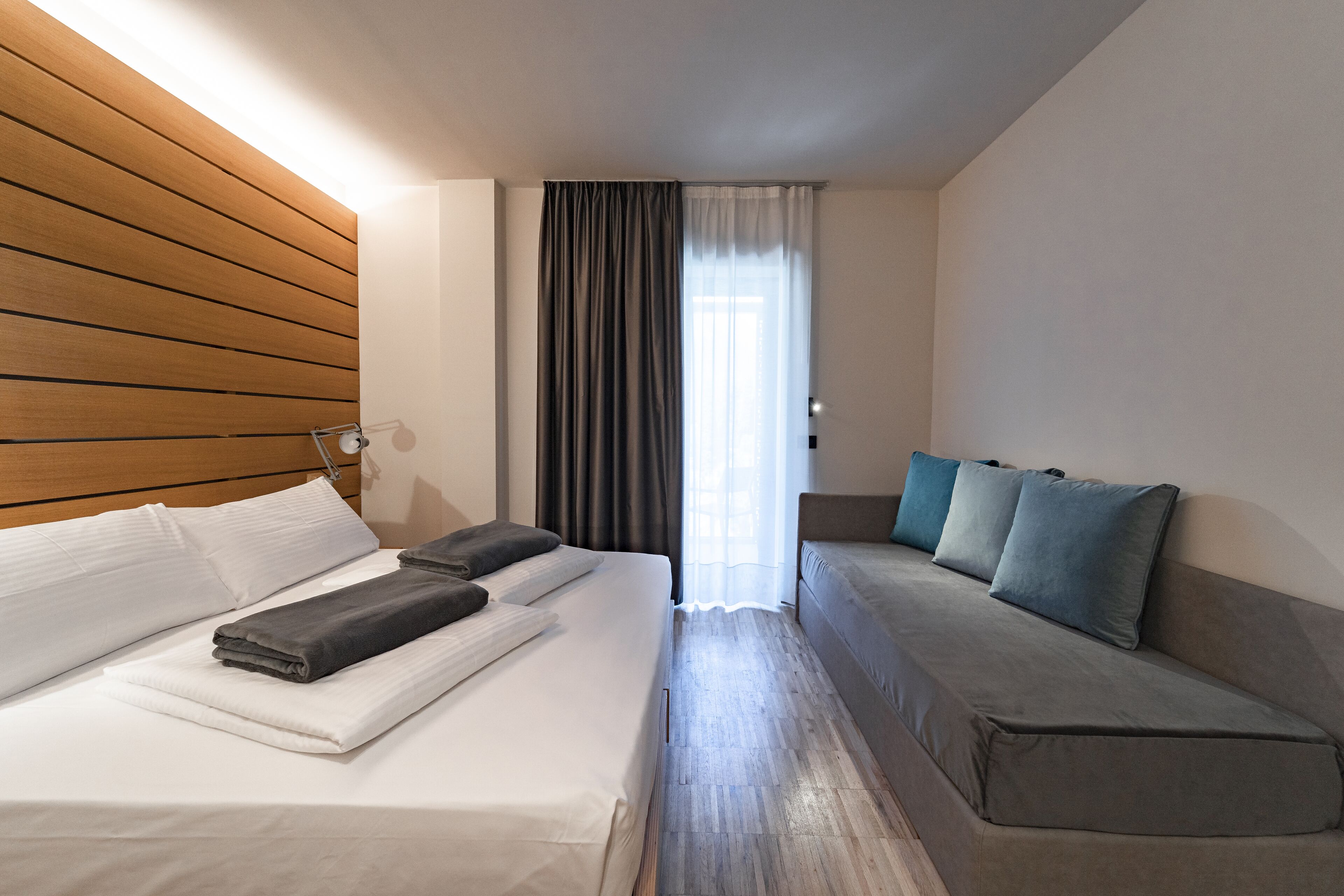 Une chambre d’hôtel avec un lit, un canapé, une tête de lit en bois et une fenêtre avec des rideaux.