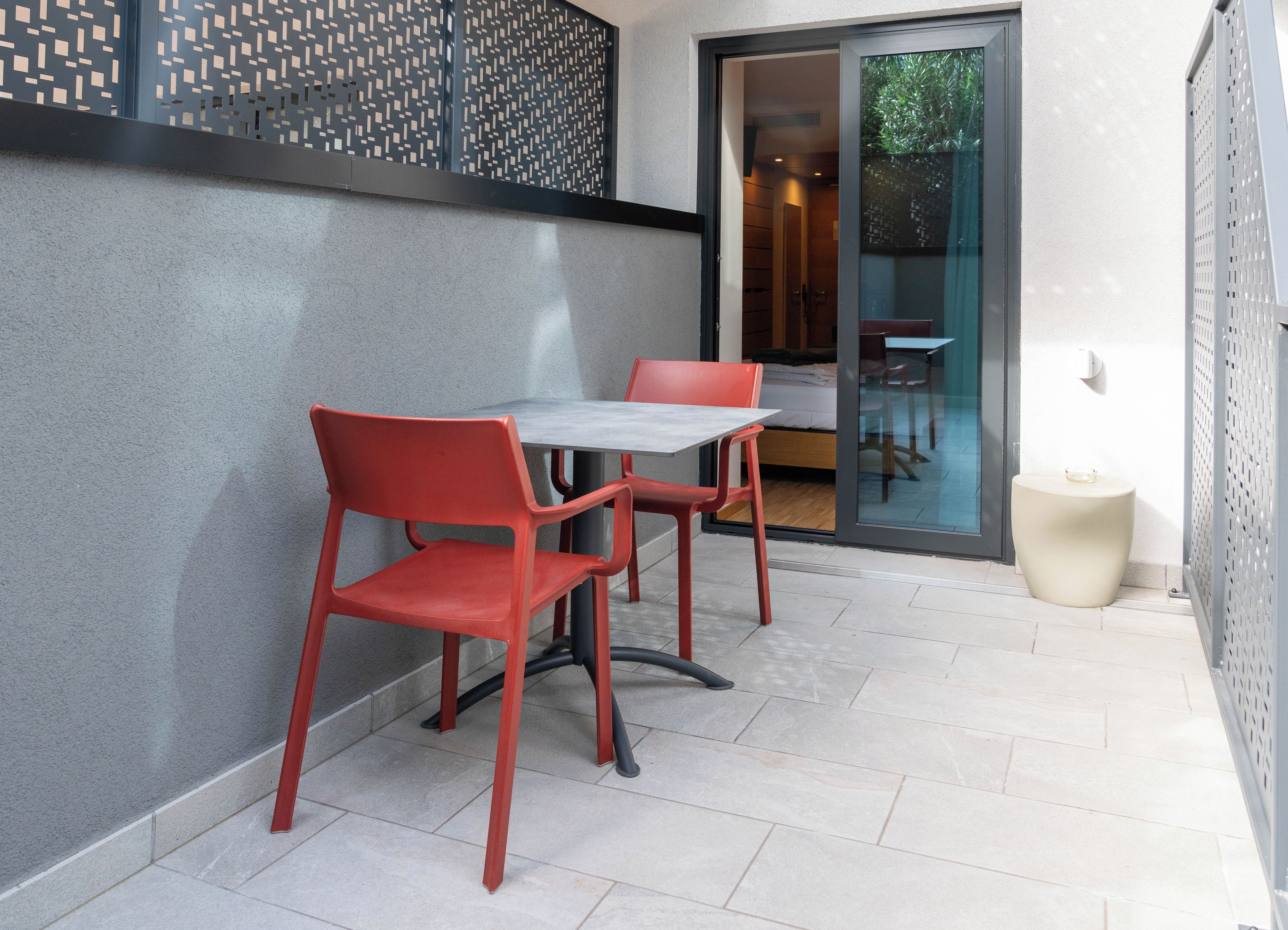 A small outdoor seating area with a table and two chairs, a sliding glass door leading to a room, and a decorative wall panel.