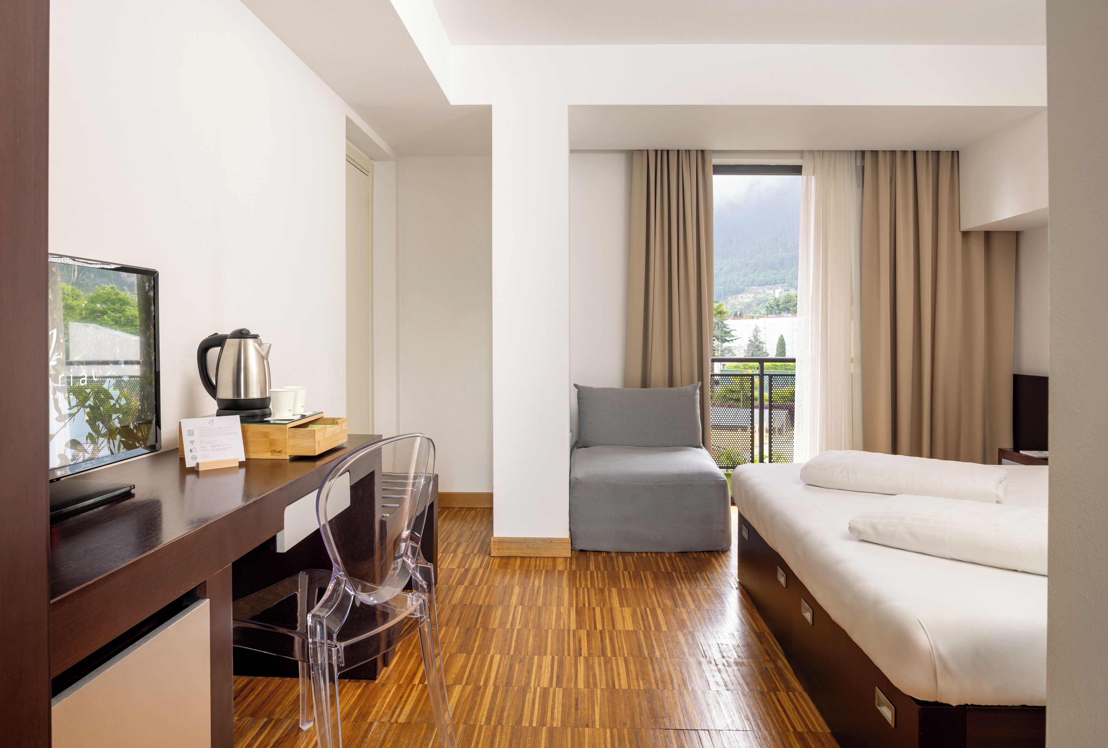 A hotel room with a wooden floor, a bed, a desk with a chair, a television, and a window with curtains.